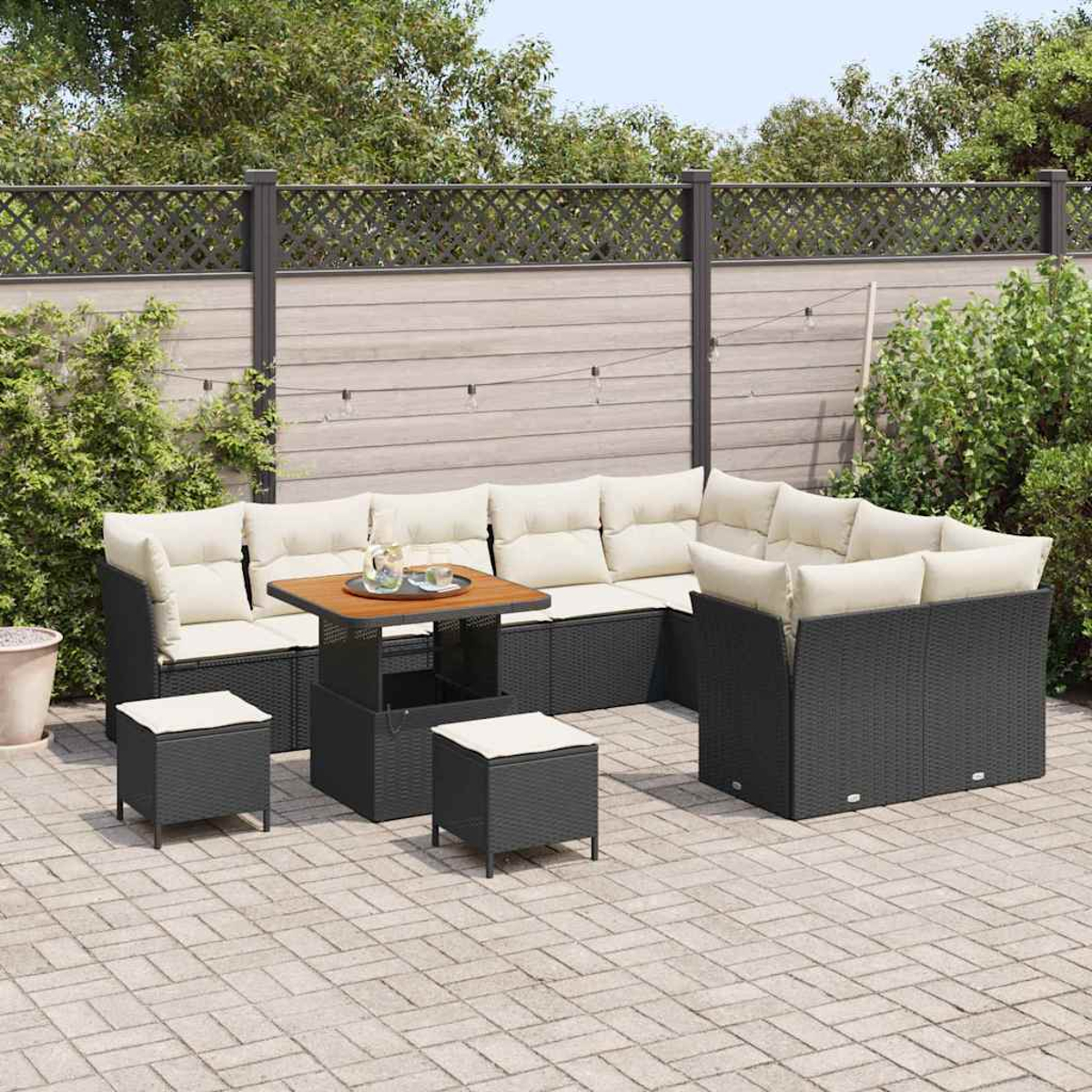 Gartenmöbel-Set mit schwarzem Korbmöbel, cremefarbenen Kissen, einem Holztisch und zwei kleinen Hockern auf einer Steinterrasse.