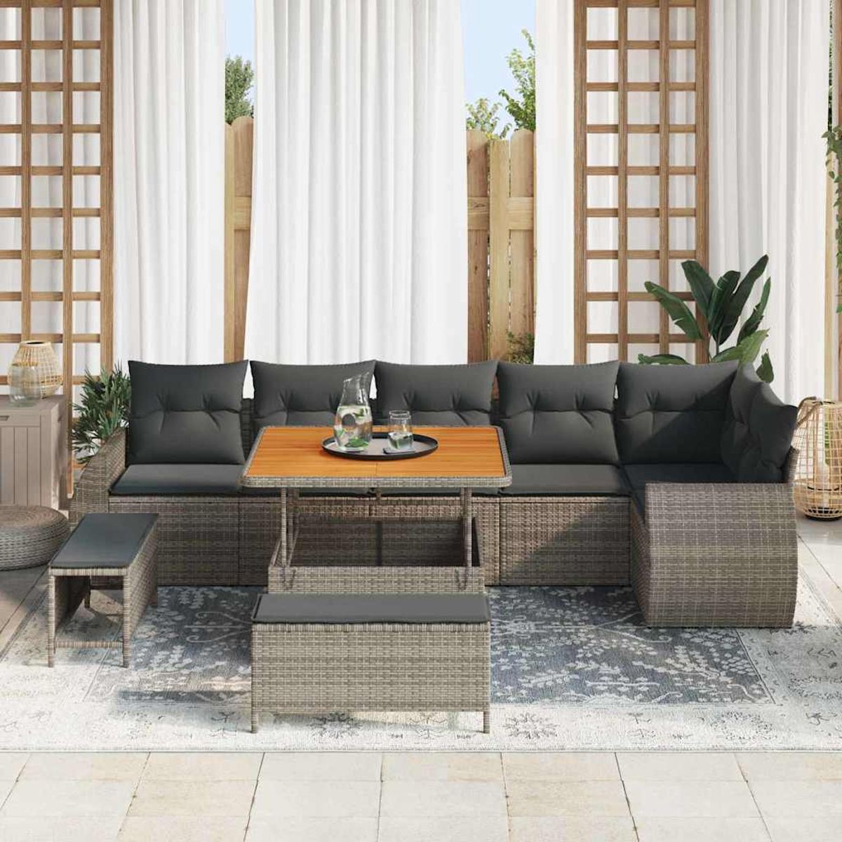 Set di mobili da esterno in vimini grigio con cuscini, tavolo in legno e panca su un tappeto fantasia, tende bianche e vista giardino.