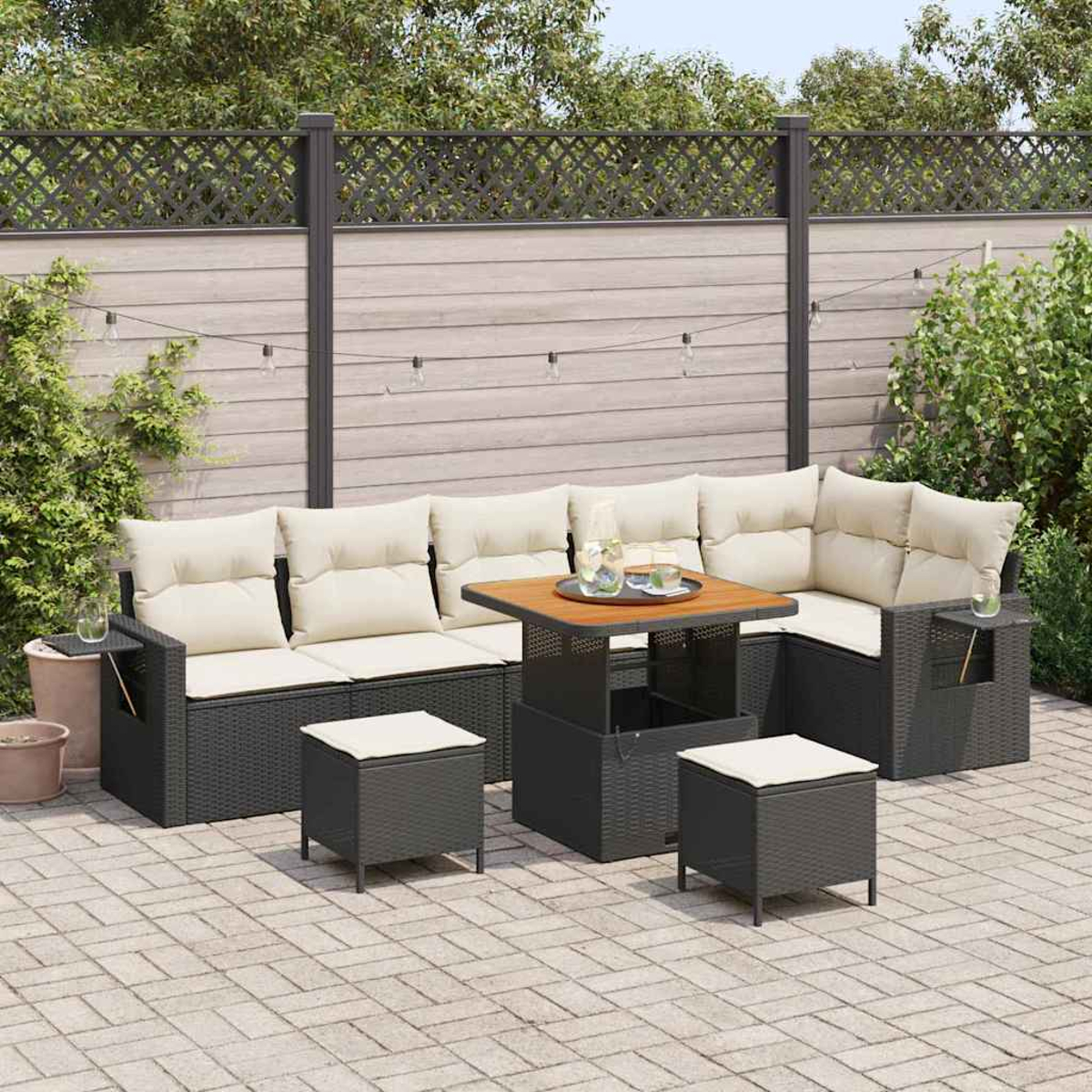 Terrassenset mit dunkelgrauen Korbmöbeln, cremefarbenen Kissen, quadratischem Tisch und zwei Hockern. Hinterhof mit Zaun und Lichterketten.