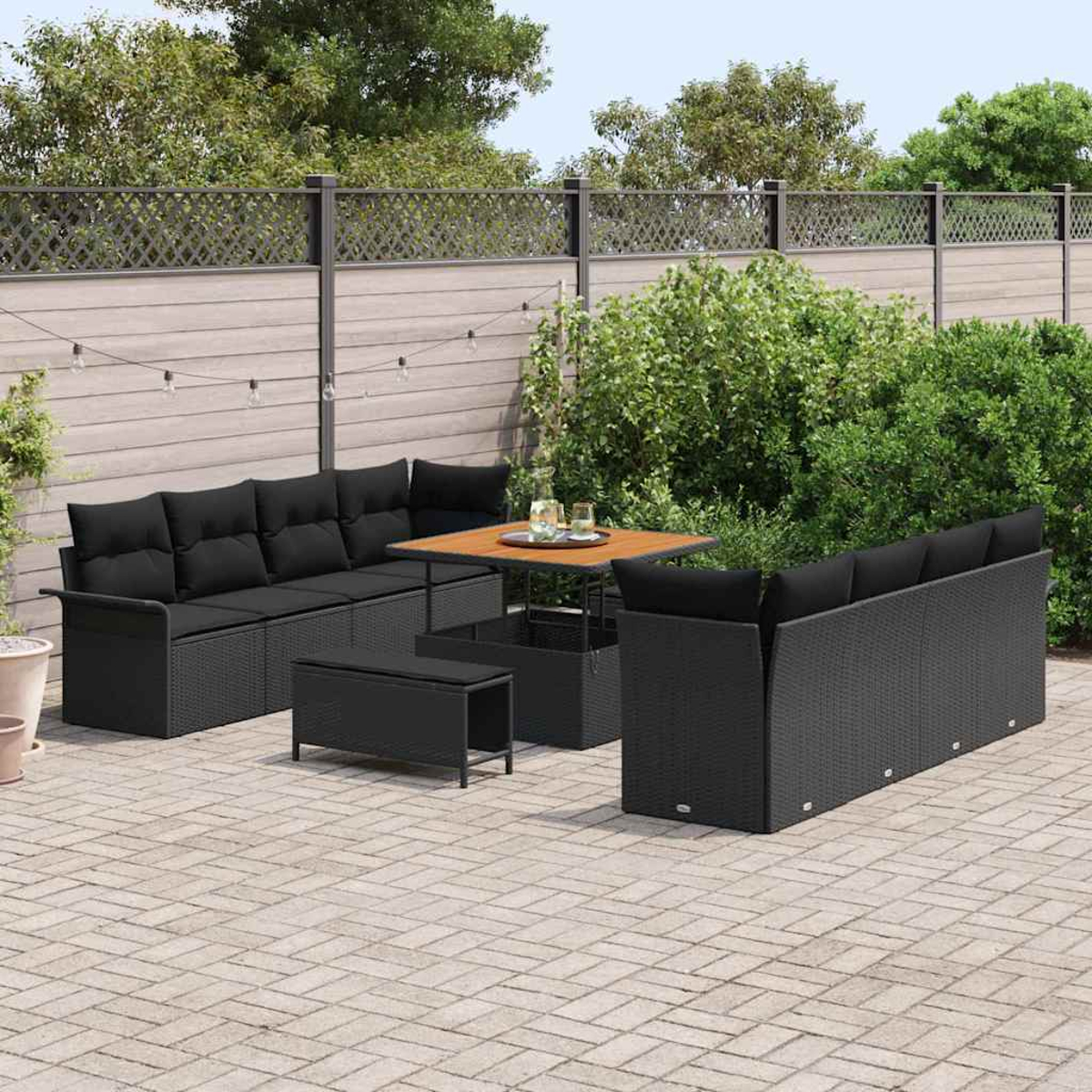Terrassenset im Freien: schwarze Korbsessel, Tisch und Bank auf einer Ziegelterrasse. Grüne Büsche und Holzzaun im Hintergrund.
