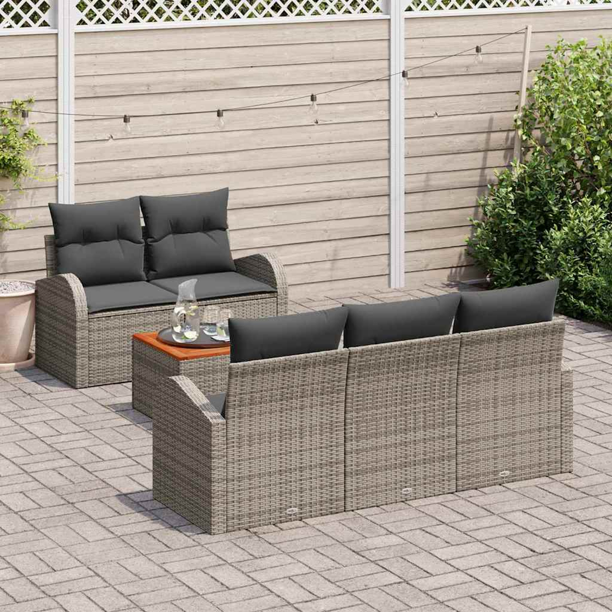 Garten-Terrassenset: graue Korbsessel mit dunkelgrauen Kissen, kleiner Holztisch mit Getränken, auf einer Steinpflasterterrasse, Holzzaun im Hintergrund.