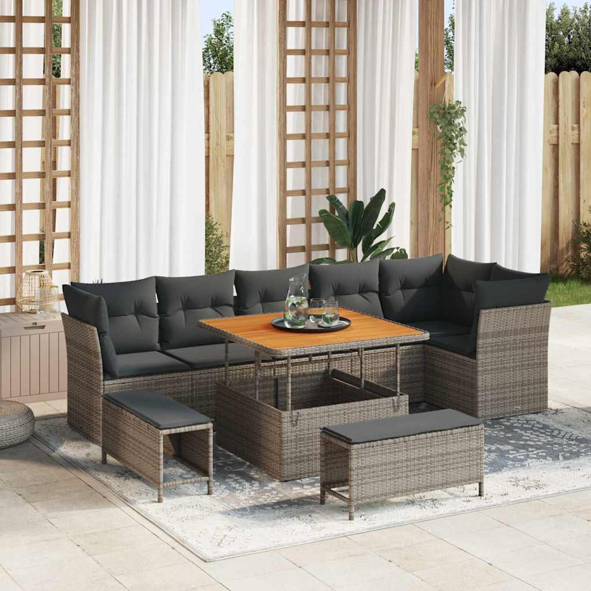 Outdoor-Rattansofa mit grauen Kissen und einem Holztisch. Zwei Bänke davor. Weiße Vorhänge und Holzzaun im Hintergrund.