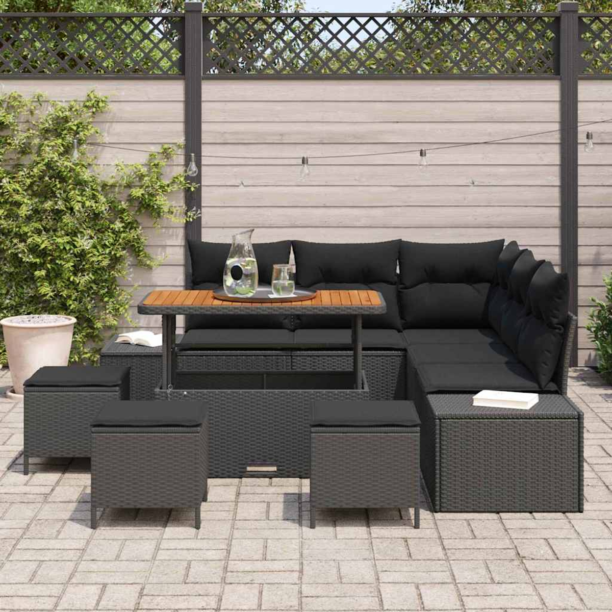 Terrassenmöbel-Set mit schwarzen Kissen, Holztisch und grauen Korbmöbeln. Auf dem Tisch steht eine Wasserkaraffe.