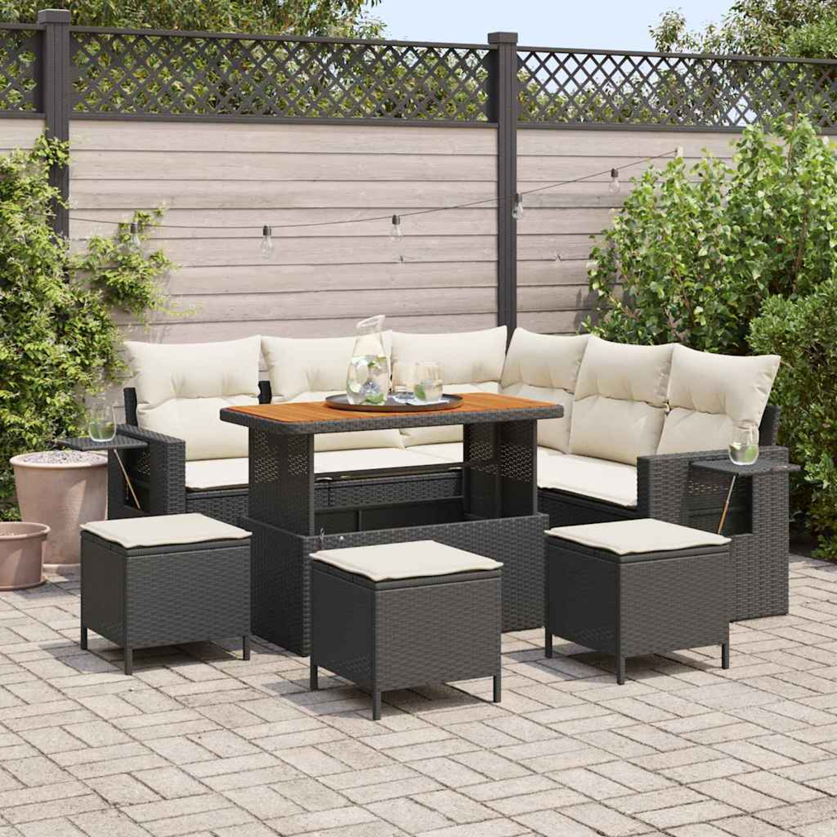 Terrassen-Set mit Sofa, Hockern und Tisch. Beige Kissen auf dunkelgrauem Korbgeflecht. Holztisch mit Getränken. Ziegel-Patio, Holzzaun.