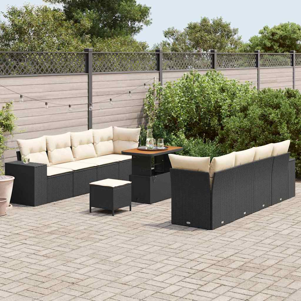 Garten-Terrassenset mit schwarzen Korbs Sofas, beigen Kissen, einem Tisch und einem kleinen Hocker auf einer Ziegel Terrasse, mit Zaun und Grün.