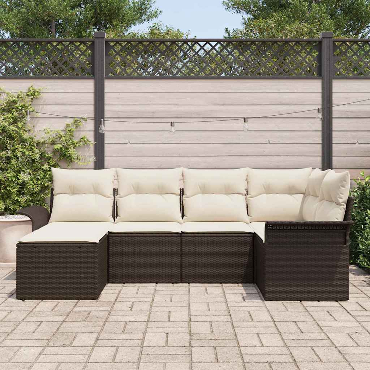Ein braunes Outdoor-Ecksofa aus Korbgeflecht mit cremefarbenen Kissen steht auf einer Terrasse. Ein Holzzaun und Grünzeug sind im Hintergrund.