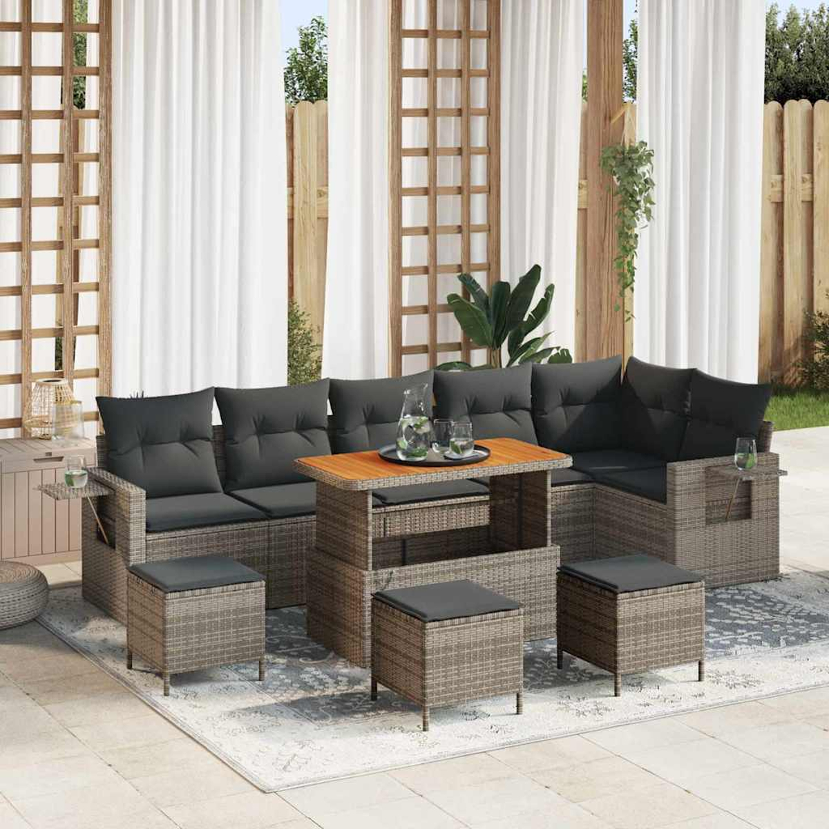 Outdoor-Rattan-Ecksofa mit grauen Kissen, Holztisch und Ottomanen. Vorhänge und Holzzaun im Hintergrund.