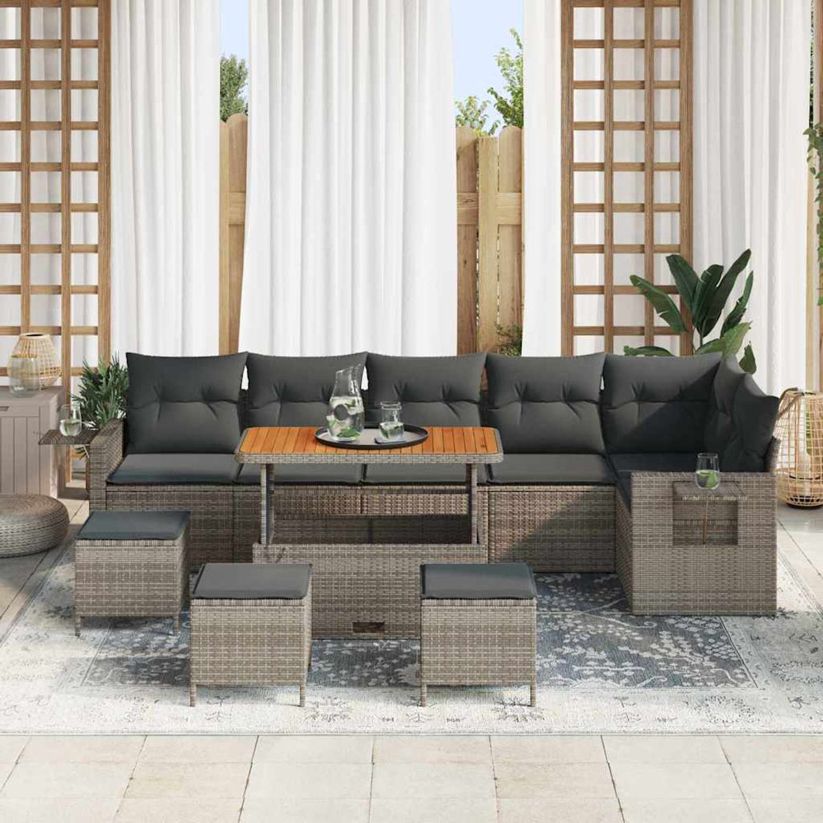Garten-Sitzgruppe mit grauen Kissen, Holztisch und Hockern auf einem gemusterten Teppich, vor weißen Vorhängen und einem Holzzaun.