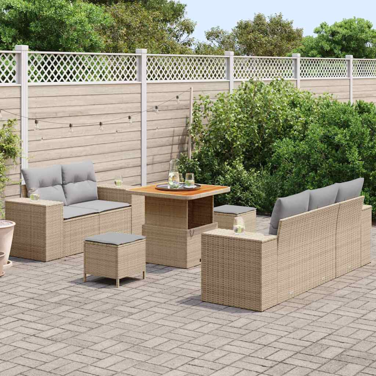 Zona de estar al aire libre con muebles beige tejidos, cojines grises, una mesa de madera y un patio de ladrillos, rodeado de vegetación y una valla.