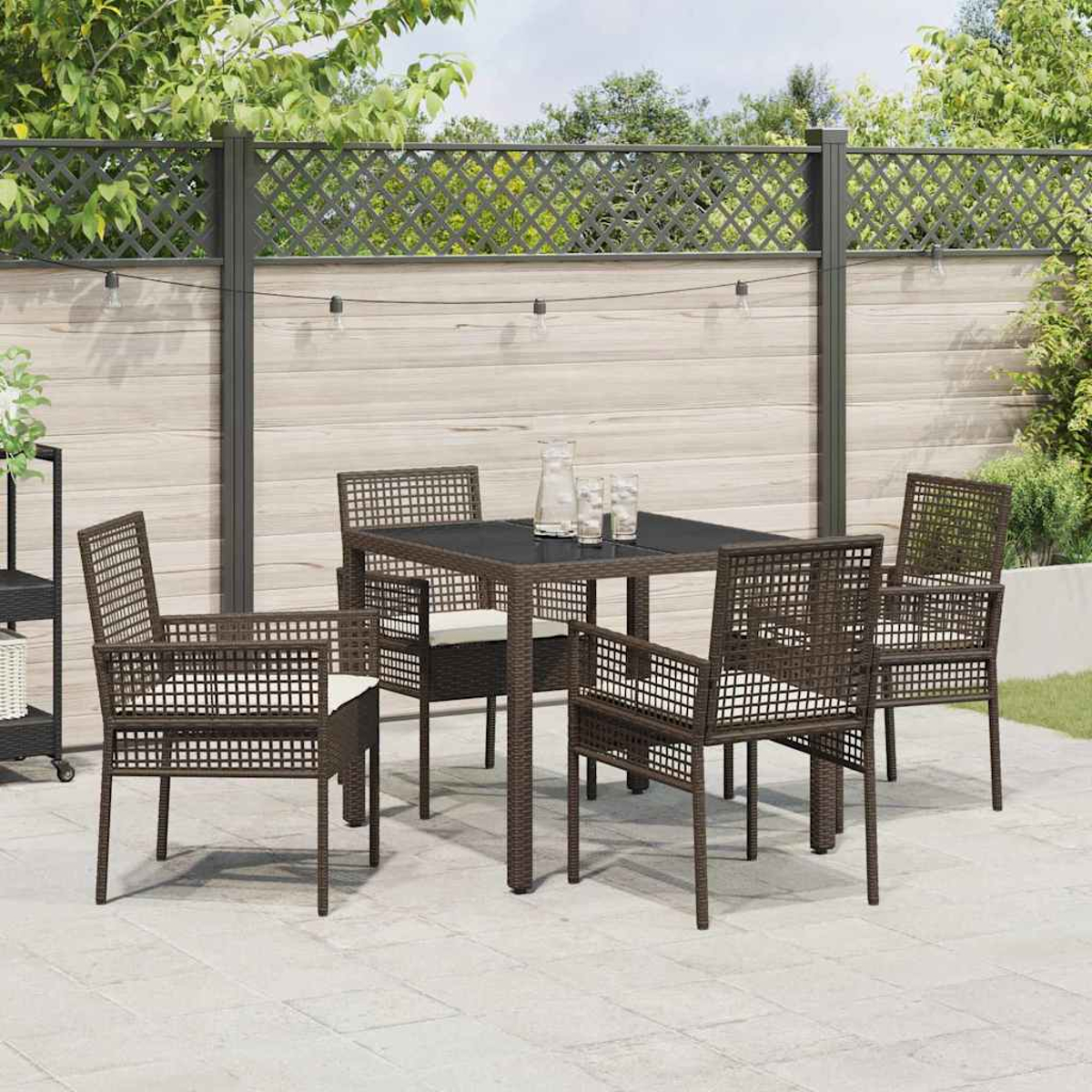 Ein Terrassenset mit Tisch und vier Stühlen. Ein Zaun und grüne Bäume sind im Hintergrund. Lichterketten sind oben.