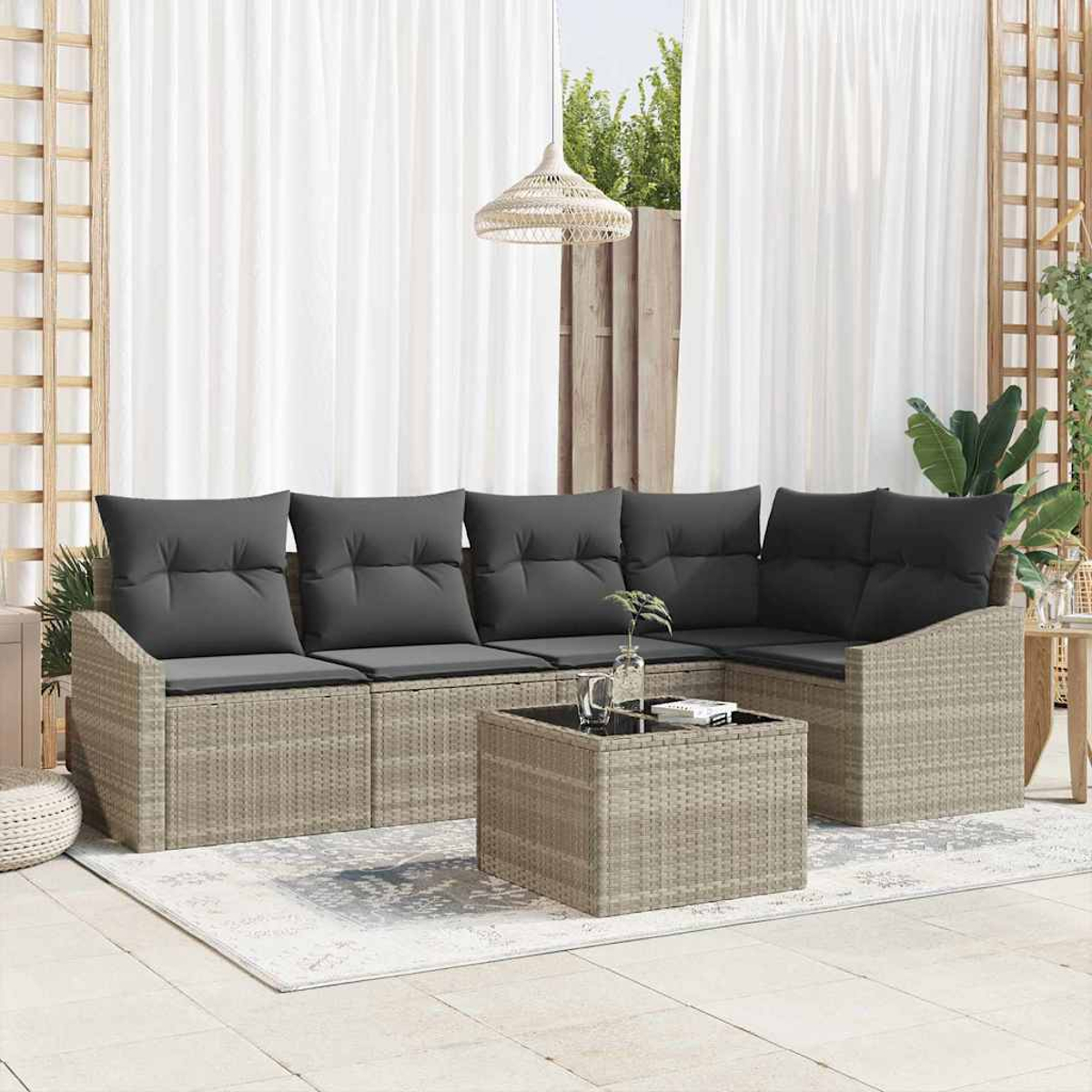 Sofá de ratán exterior con cojines grises, mesa de centro, cortinas blancas y una lámpara colgante. La escena es en un patio.