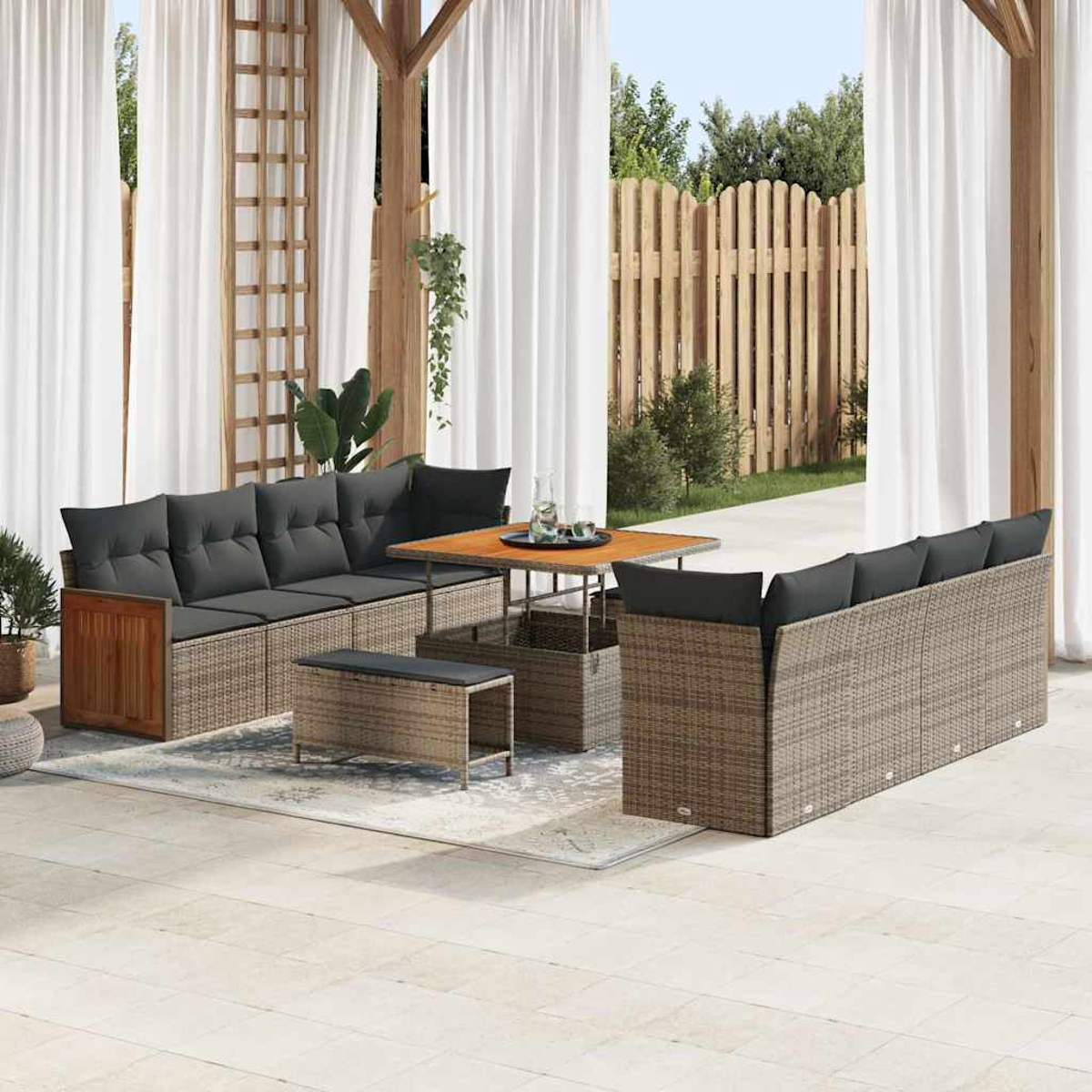 Outdoor-Sitzbereich mit Sofa, Tisch und Bank. Graue Kissen auf gewebten Möbeln. Weiße Vorhänge und ein Holzzaun im Hintergrund.