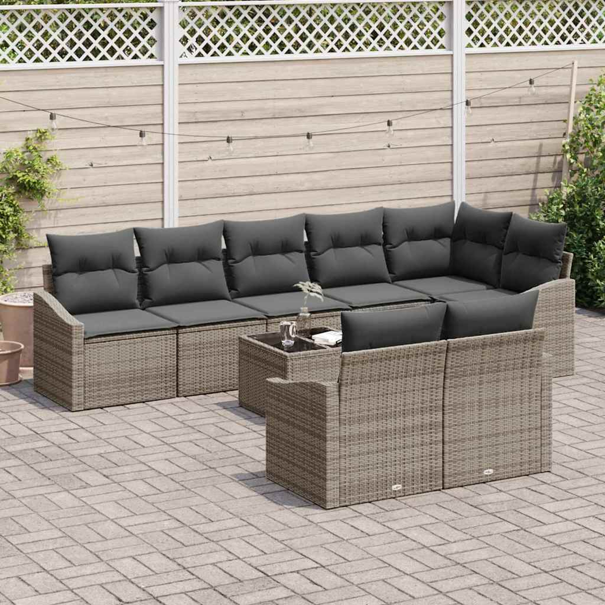 Rattan-Sofa-Set im Freien mit grauen Kissen auf einer Ziegelterrasse. Im Hintergrund ein Holzzaun und dekorative Lichter.