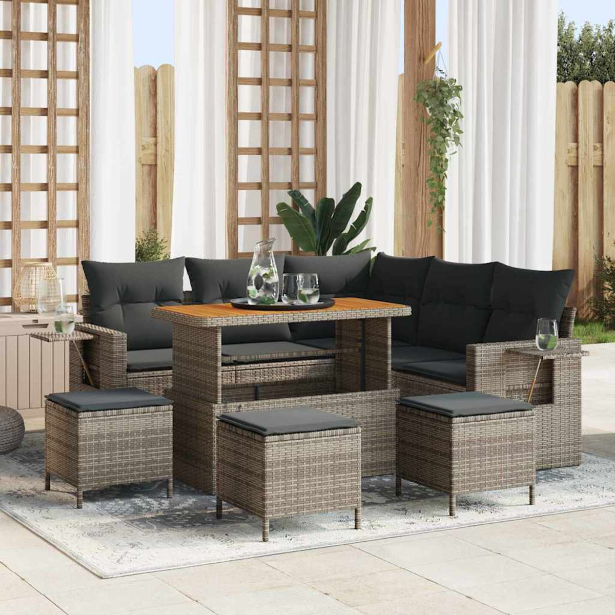 Garten-Sitzgruppe mit Tisch, Sofa und Ottomanen. Graue Kissen auf gewebten Möbeln. Weiße Vorhänge und hölzerner Spalier im Hintergrund.