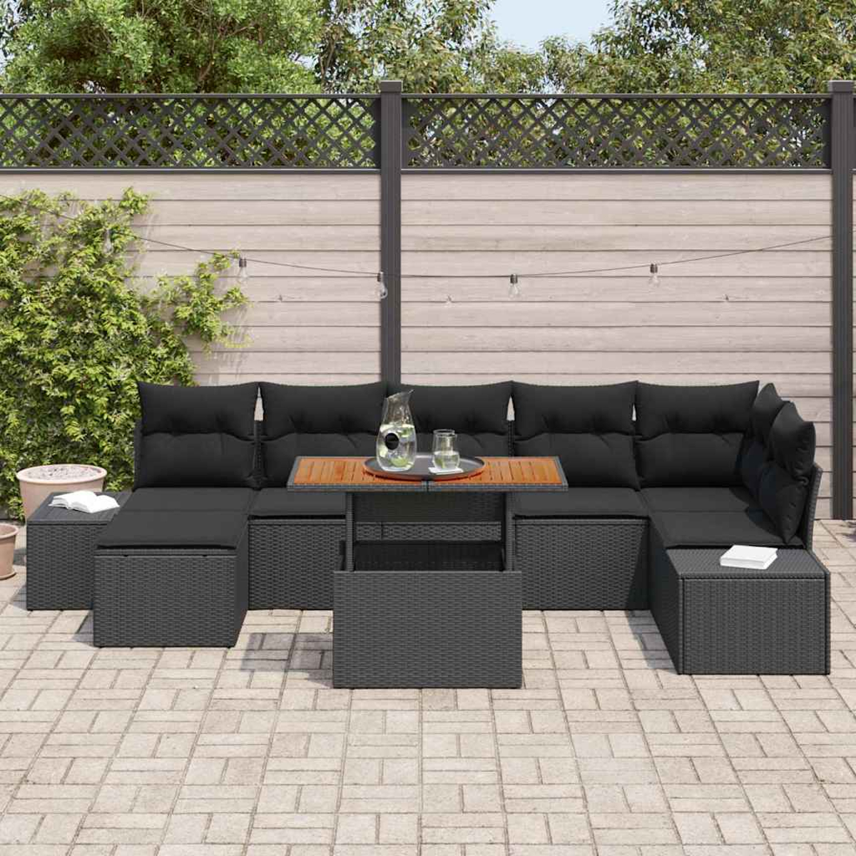 Garten-Terrassenset mit schwarzen Kissen, einem Holztisch und grauen Korbmöbeln. Im Hintergrund ein Holzzaun und Grün.