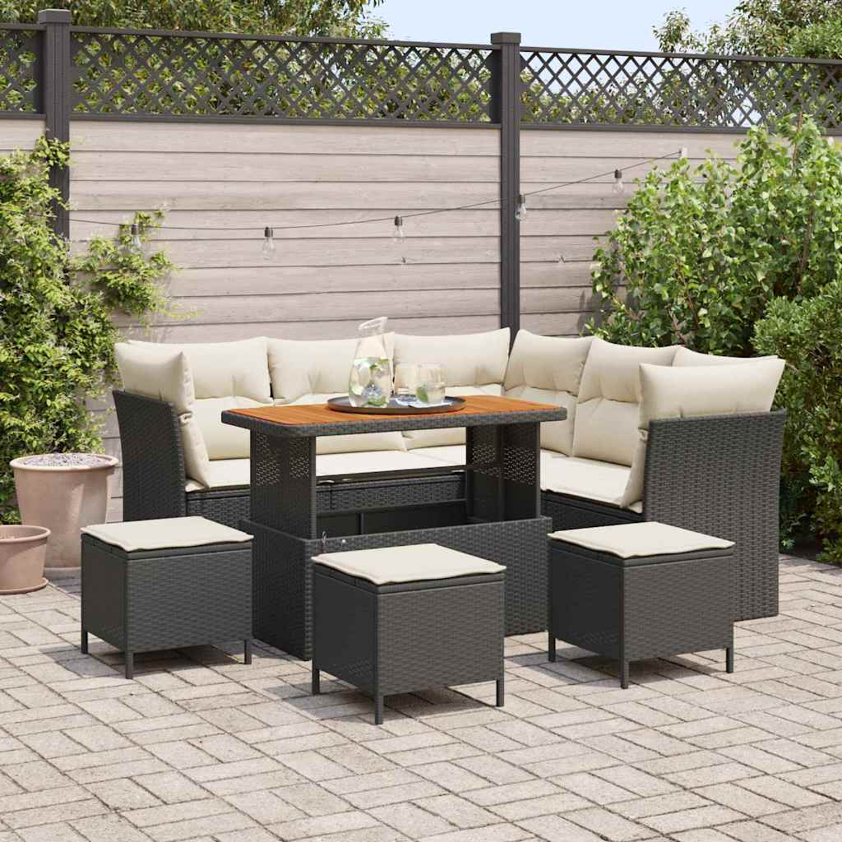 Set divano angolare da esterno con pouf e tavolino su un patio in mattoni. Cuscini color crema, vimini grigio scuro e tavolo in legno.