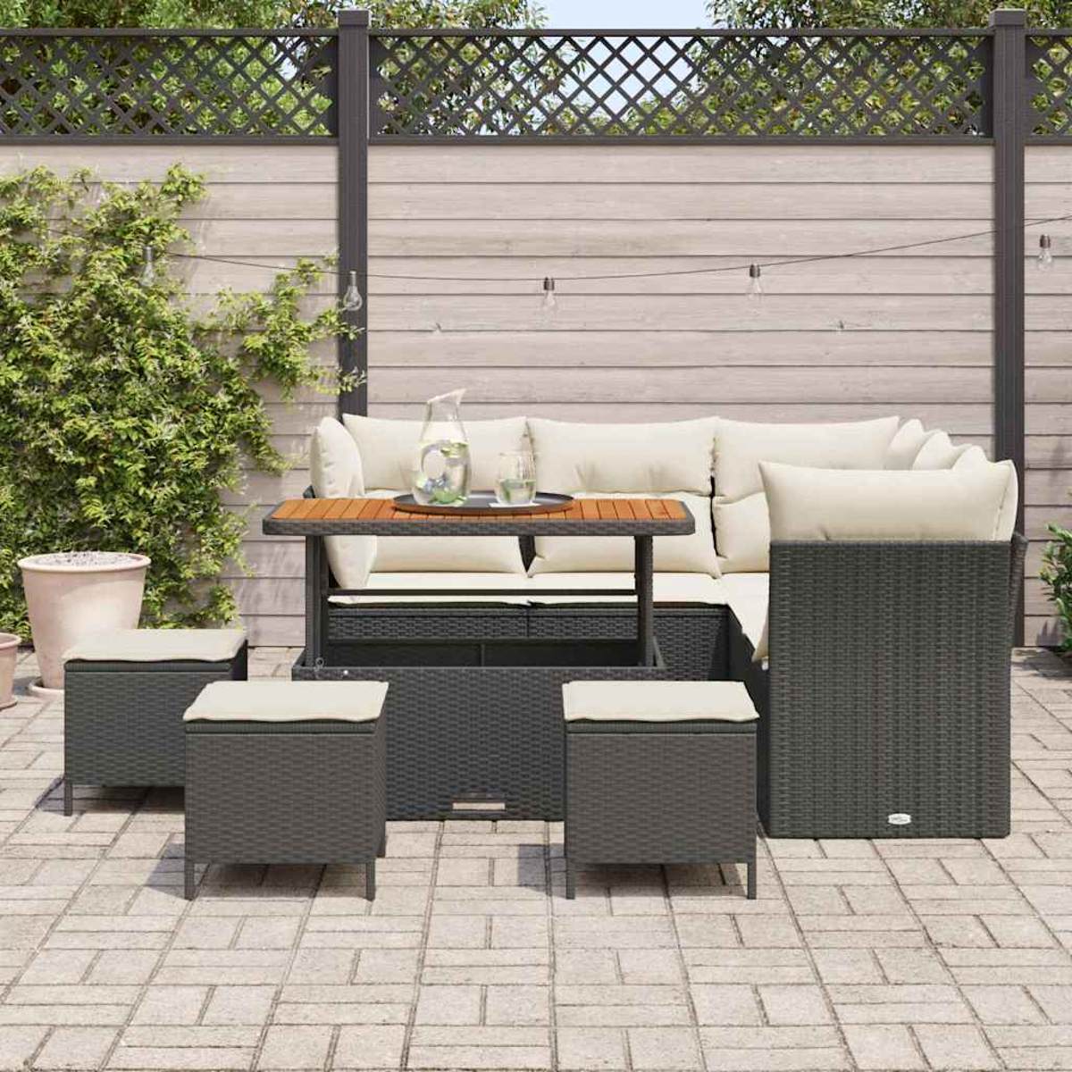 Set di sedute per esterni con tavolo, cuscini e sgabelli. Il set si trova su un patio con una staccionata in legno e piante sullo sfondo.