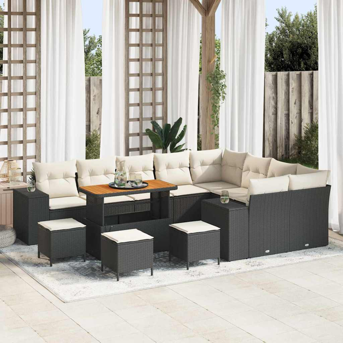Set da patio all'aperto con divano in vimini grigio scuro, cuscini beige, tavolo in legno e piccoli pouf su un tappeto. Tende bianche sullo sfondo.