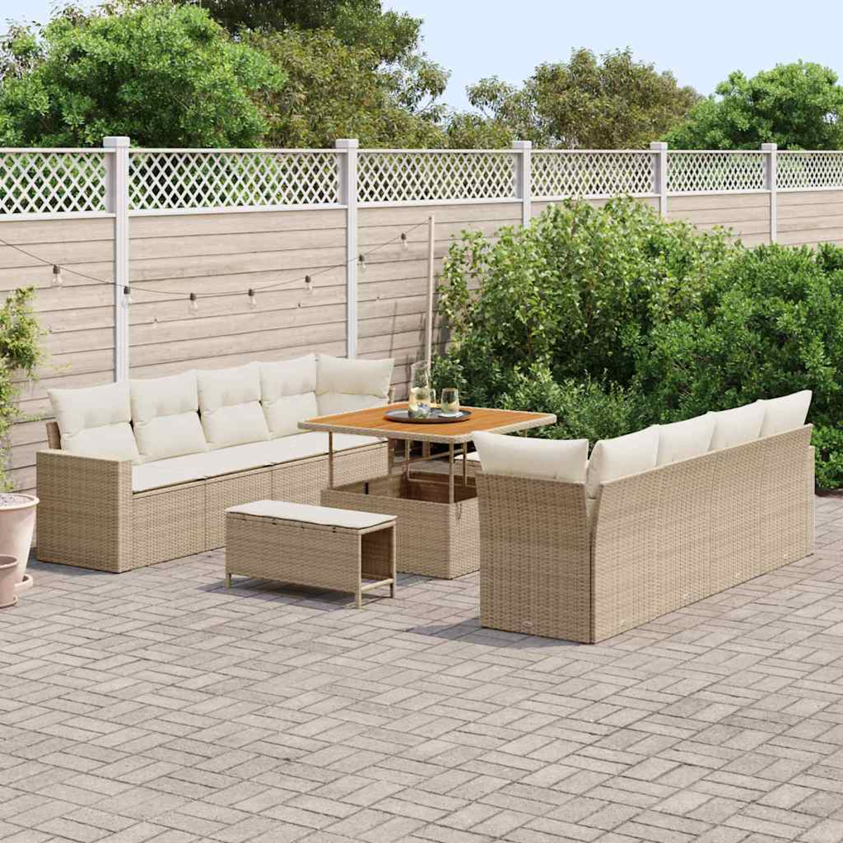 Terrassenset im Freien: beige Korbsessel, Tisch und Bank auf einer Ziegelpflasterung. Weißer Gitterzaun und grüne Büsche im Hintergrund.