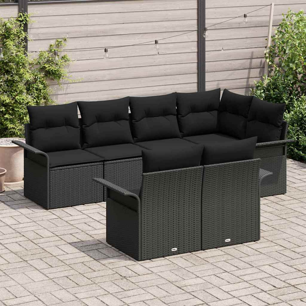 Outdoor-Ecksofa mit schwarzen Kissen, grauem Korbrahmen und zwei Sesseln auf einer Backstein-Terrasse, mit Pflanzen im Hintergrund.