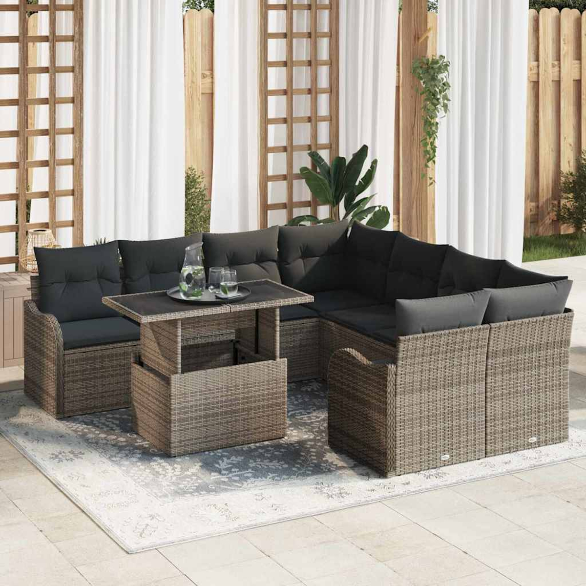 Juego de sofás de mimbre para exteriores con cojines negros y una mesa de centro elevable sobre una alfombra estampada, contra un enrejado de madera y cortinas blancas.