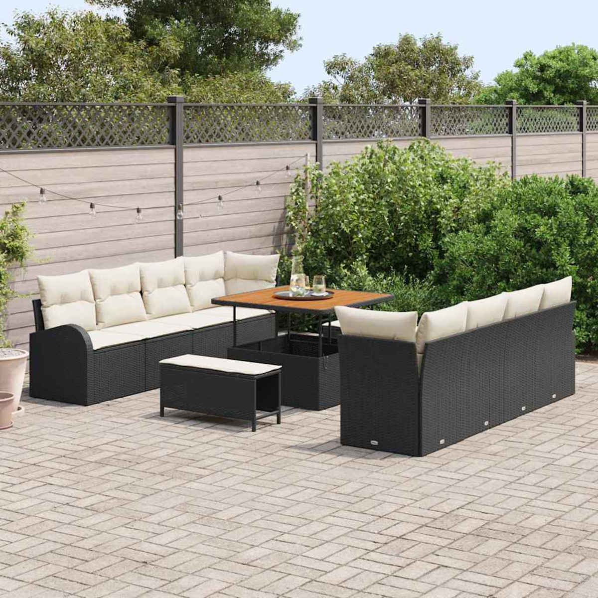 Garten-Set. Schwarze Korbmöbel mit cremefarbenen Kissen. Ein Tisch und eine Bank sind ebenfalls vorhanden. Grüne Büsche und ein Zaun im Hintergrund.