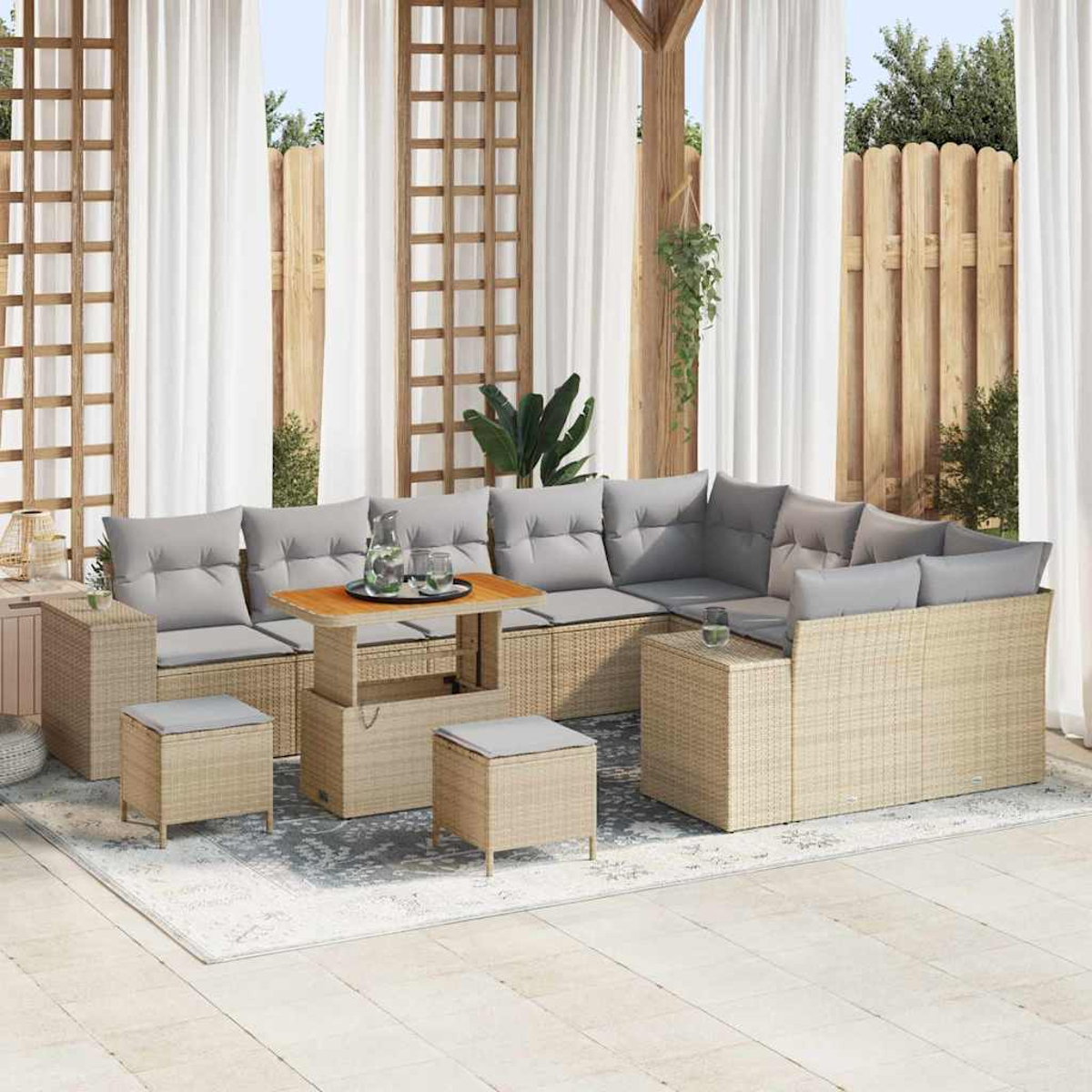 Set divano in rattan da esterno con cuscini grigi, tavolo in legno e due sgabelli su un tappeto fantasia. Tende bianche e recinzione in legno.