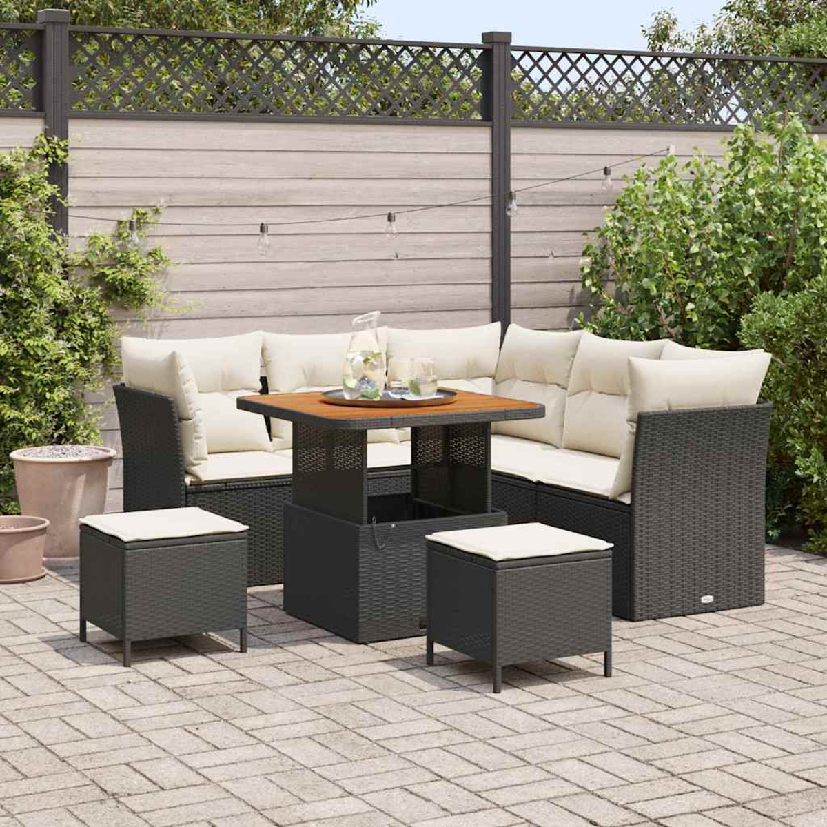 Set da patio all'aperto con mobili in vimini scuro, cuscini beige e un tavolo in legno. Include due pouf. Contro una recinzione e vegetazione.