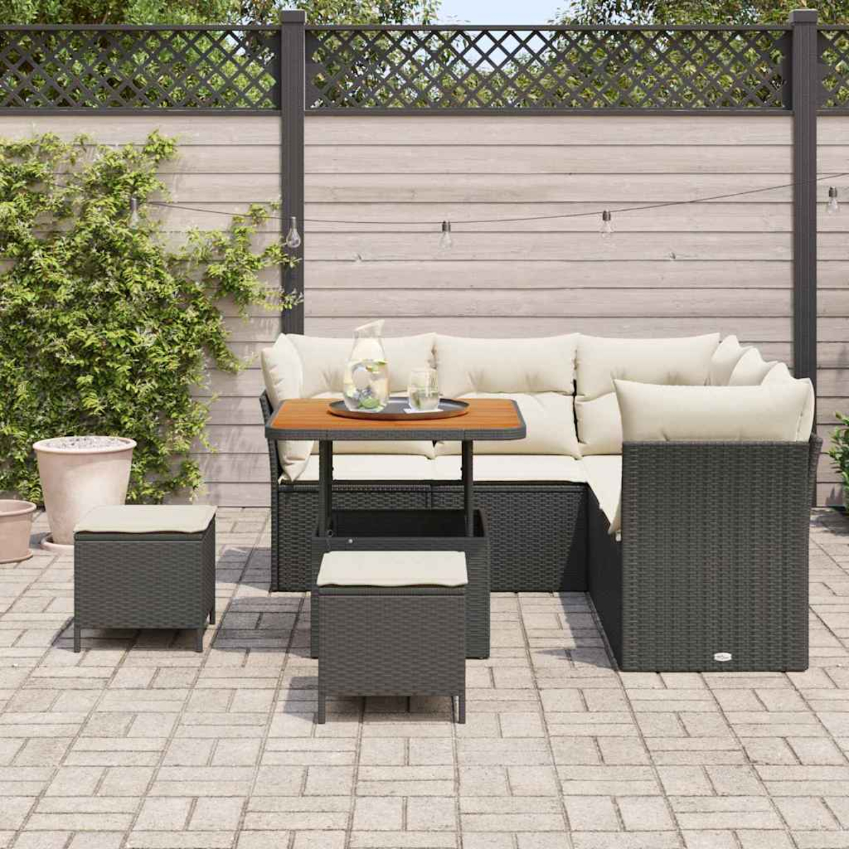 Set patio esterno con mobili in vimini grigi, cuscini color crema e un tavolo in legno. Una recinzione e piante in vaso sullo sfondo.