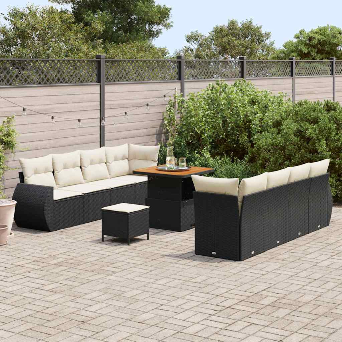 Terrassen-Set mit schwarzen Korbsesseln und Tisch mit cremefarbenen Kissen. Ein Holzzaun und üppiges Grün im Hintergrund.