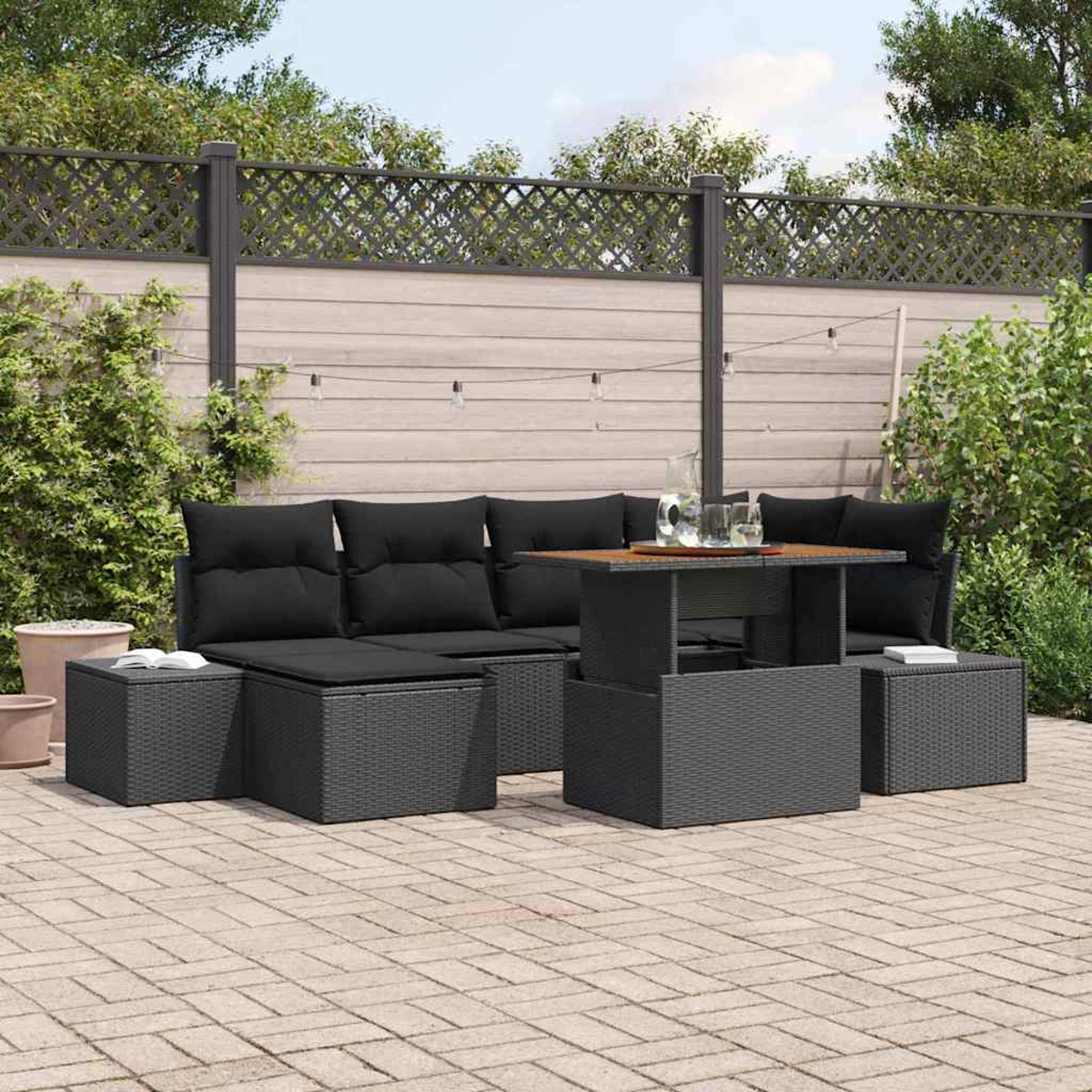 Schwarzes Outdoor-Rattan-Möbelset mit Kissen, Tisch und Ottomanen auf einer Terrasse. Hintergrund: ein Zaun mit Pflanzen.