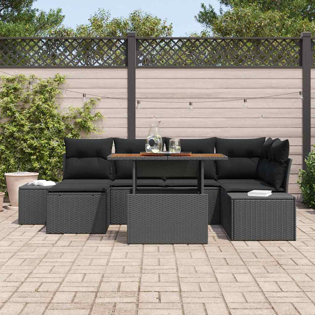 Terrassenset im Freien mit grauen Korbmöbeln, schwarzen Kissen und einem Holztisch. Auf dem Tisch stehen ein Krug und Gläser.
