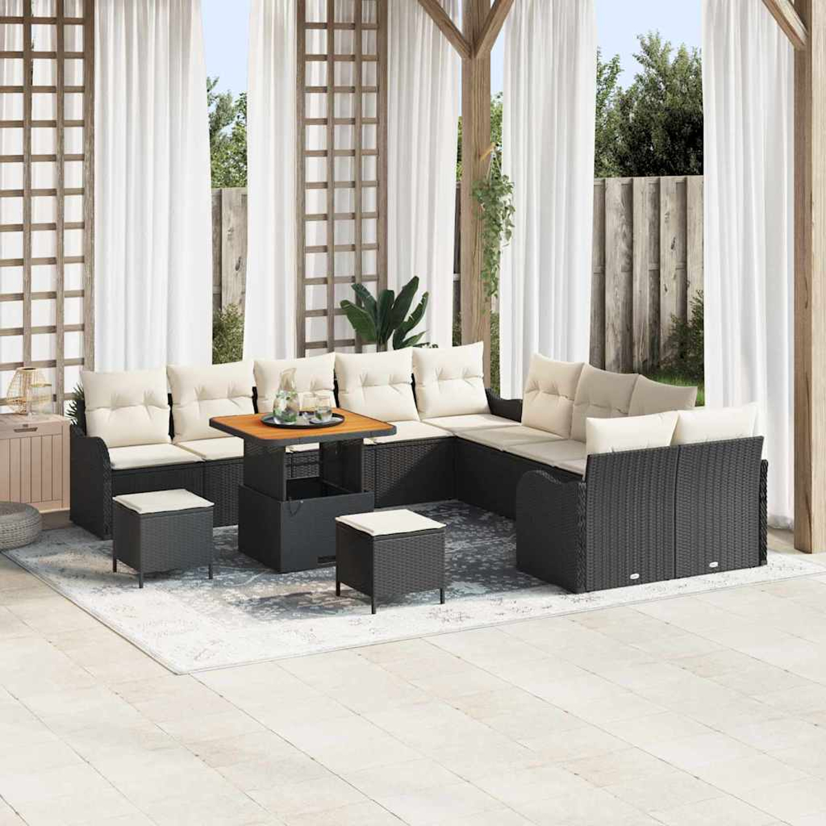 Outdoor-Sitzbereich mit schwarzen Korbmöbeln, cremefarbenen Kissen, einem Holztisch und Hockern. Weiße Vorhänge und ein Teppich vervollständigen die Szene.
