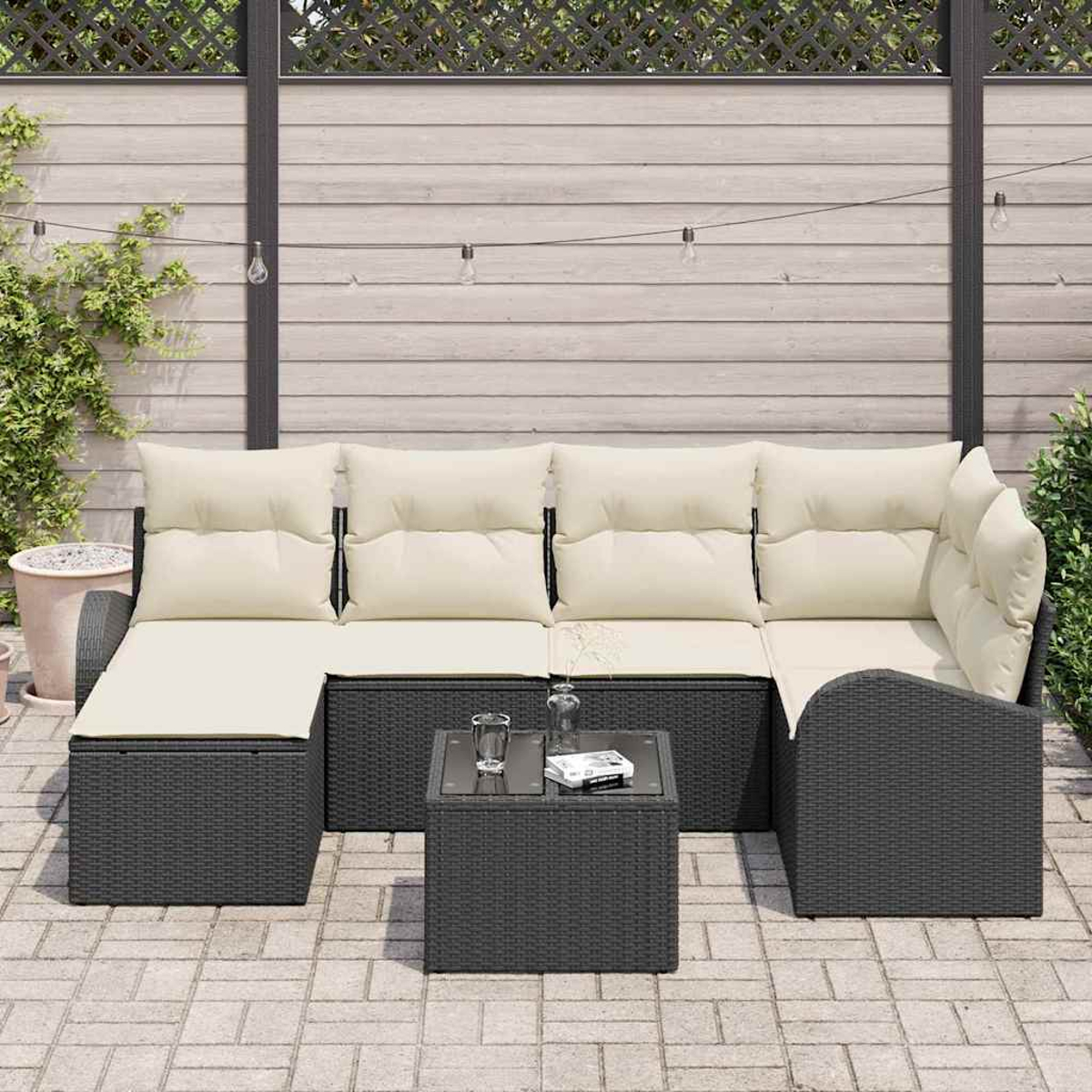 Ein dunkelgraues Outdoor-Ecksofa aus Korbgeflecht mit cremefarbenen Kissen, einem kleinen Tisch und einem Holzzaun im Hintergrund.