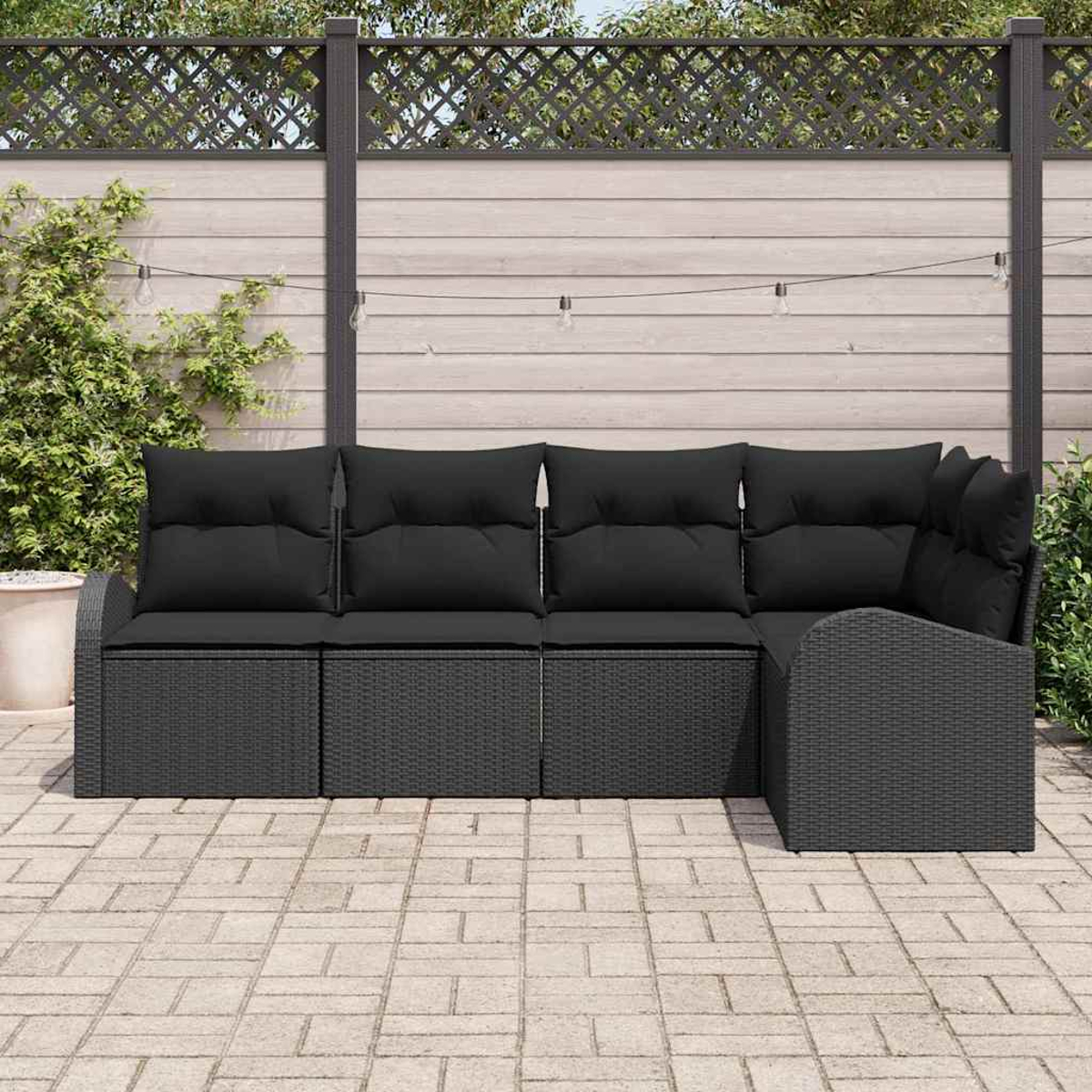 Ein schwarzes Outdoor-Ecksofa aus Korbgeflecht mit schwarzen Kissen steht auf einer Backstein-Terrasse. Im Hintergrund ein Holzzaun mit Gitteraufsatz.