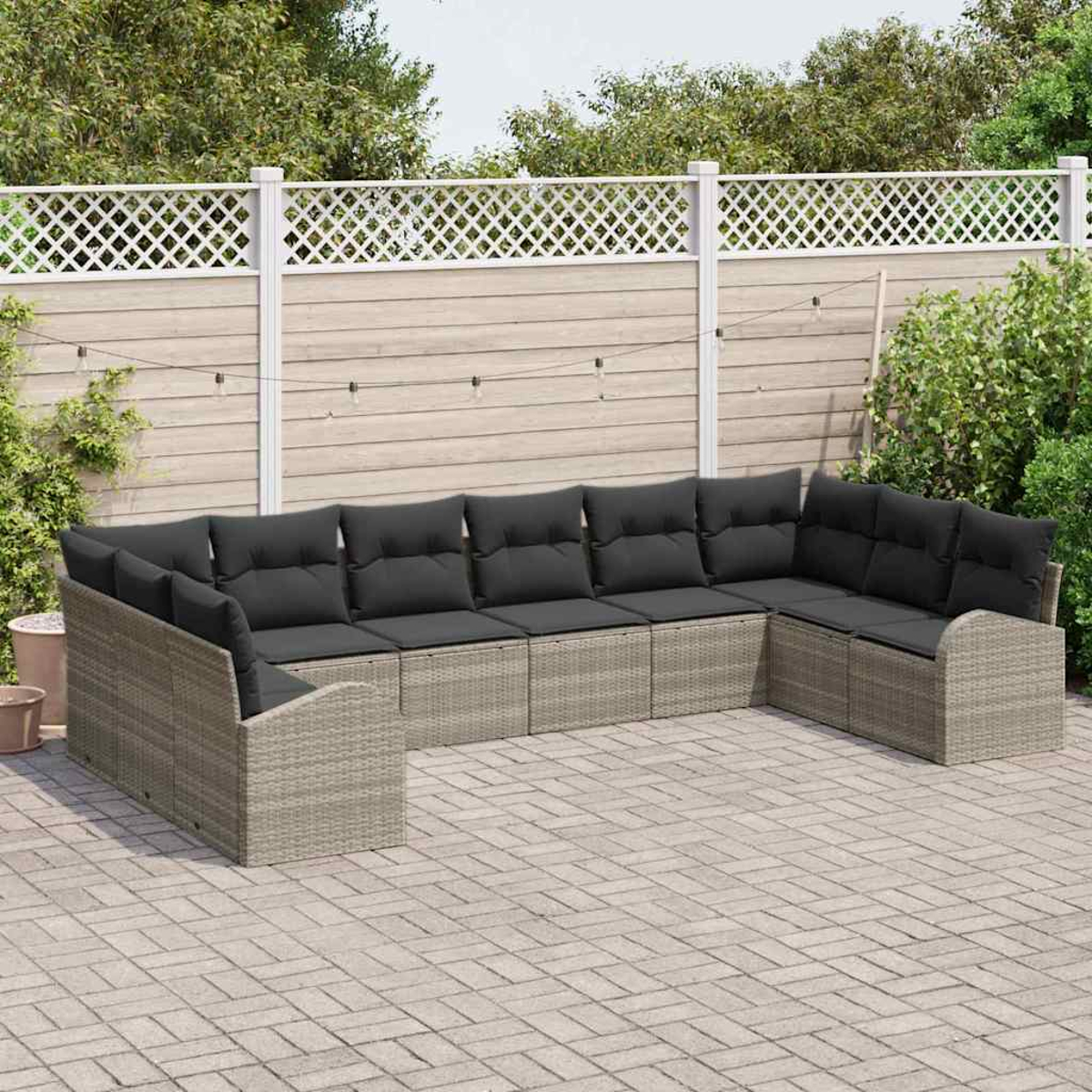 Ein graues Rattan-Ecksofa mit schwarzen Kissen steht auf einer Backstein-Terrasse. Ein weißer Gitterzaun und Grünzeug sind im Hintergrund.
