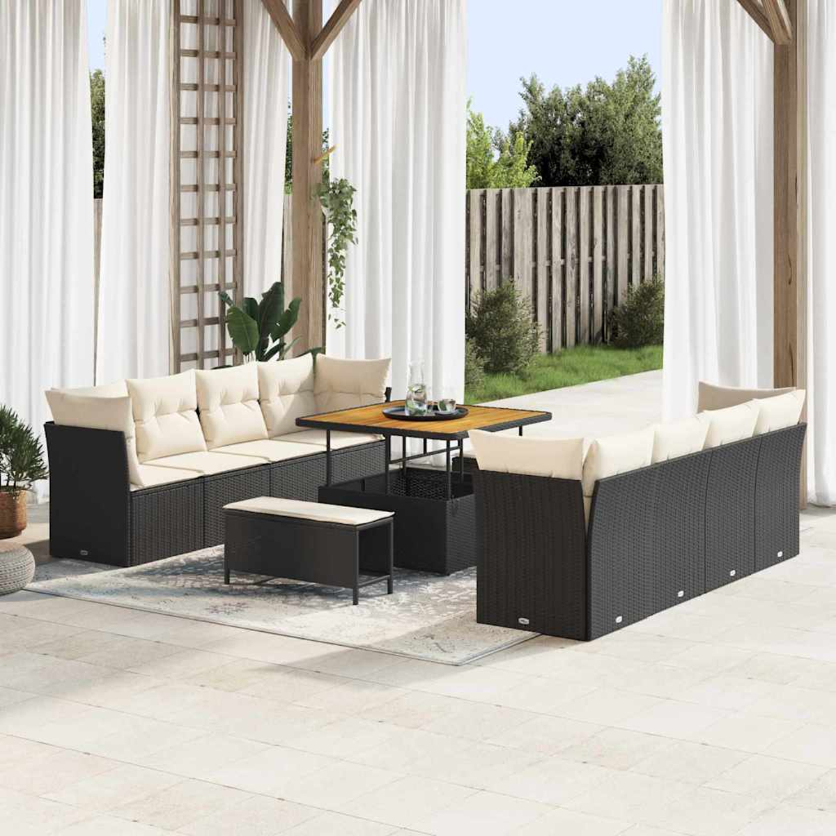 Un juego de patio al aire libre con muebles de mimbre negro, cojines crema y una mesa de madera. Cortinas blancas y una valla de madera al fondo.