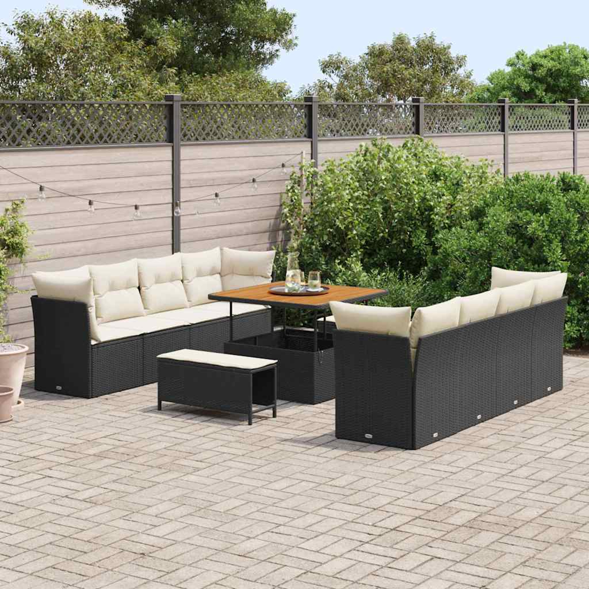 Juego de patio al aire libre. Sofás de mimbre negro con cojines crema, mesa de madera y banco en un patio de ladrillos. Arbustos verdes y valla al fondo.