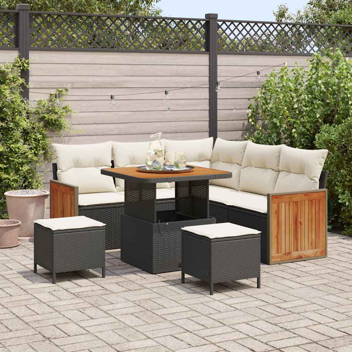 Garten-Sitzgruppe mit Tisch, Sofa und zwei Hockern. Das Set befindet sich auf einer Terrasse mit Holzzaun und Grün im Hintergrund.