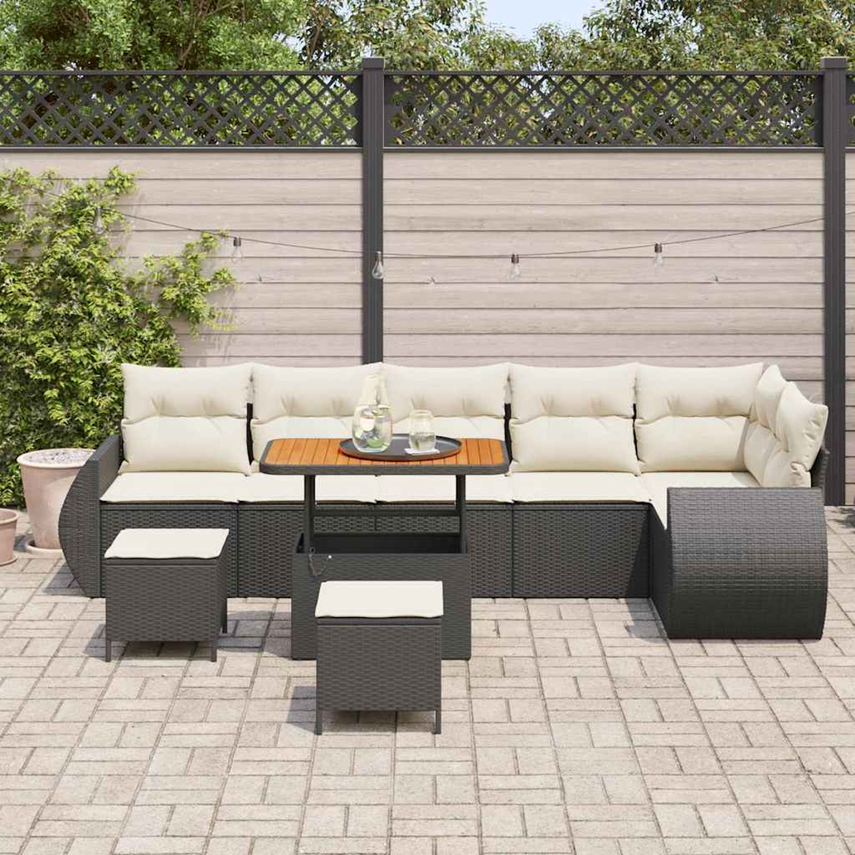 Terrassengarnitur mit Sofa, kleinem Tisch und Hockern. Cremefarbene Kissen auf dunkelgrauen Korbmöbeln. Holzzaun und Pflanzen im Hintergrund.