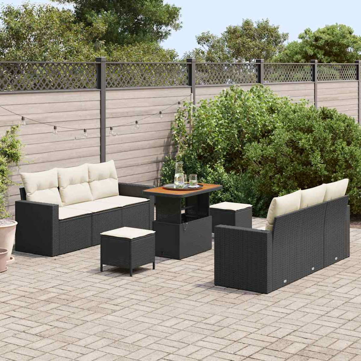 Garten-Terrassenset mit dunklen Korbmöbeln, cremefarbenen Kissen, einem Tisch und zwei Ottomanen auf einer Backsteinterrasse, mit Zaun und Grün.