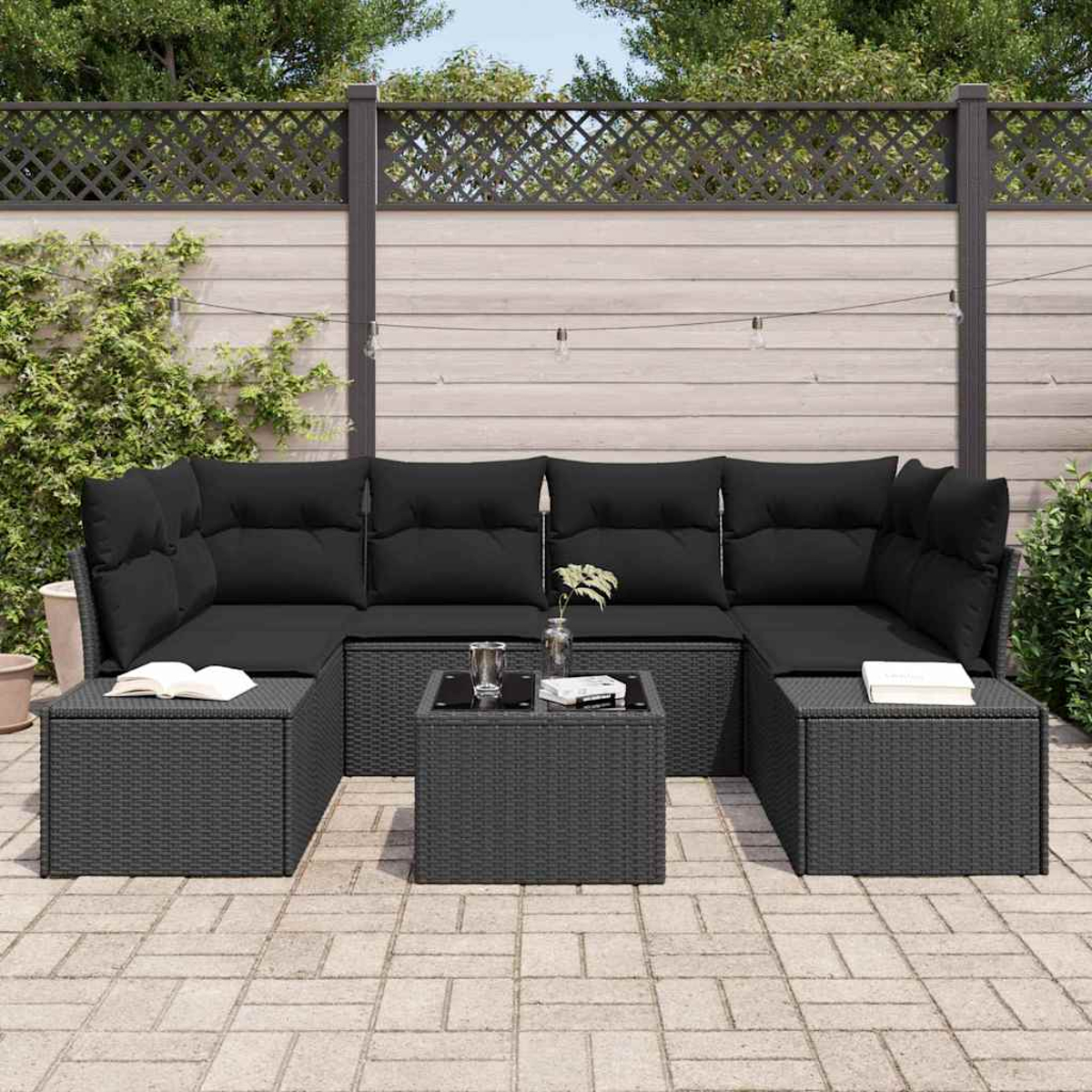 Un juego de patio negro con cojines, dos otomanas y una mesa de centro. Libros y plantas están en la mesa. Una valla de madera está en el fondo.