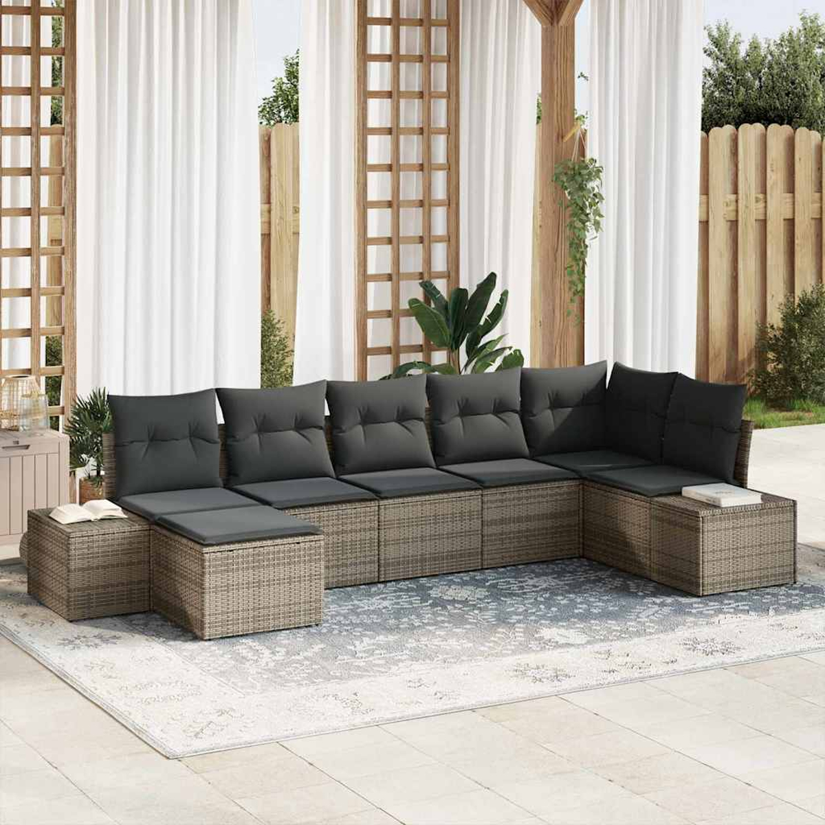 Ein Sitzbereich im Freien mit einem grauen Ecksofa auf einem gemusterten Teppich. Weiße Vorhänge und Holzstrukturen befinden sich im Hintergrund.