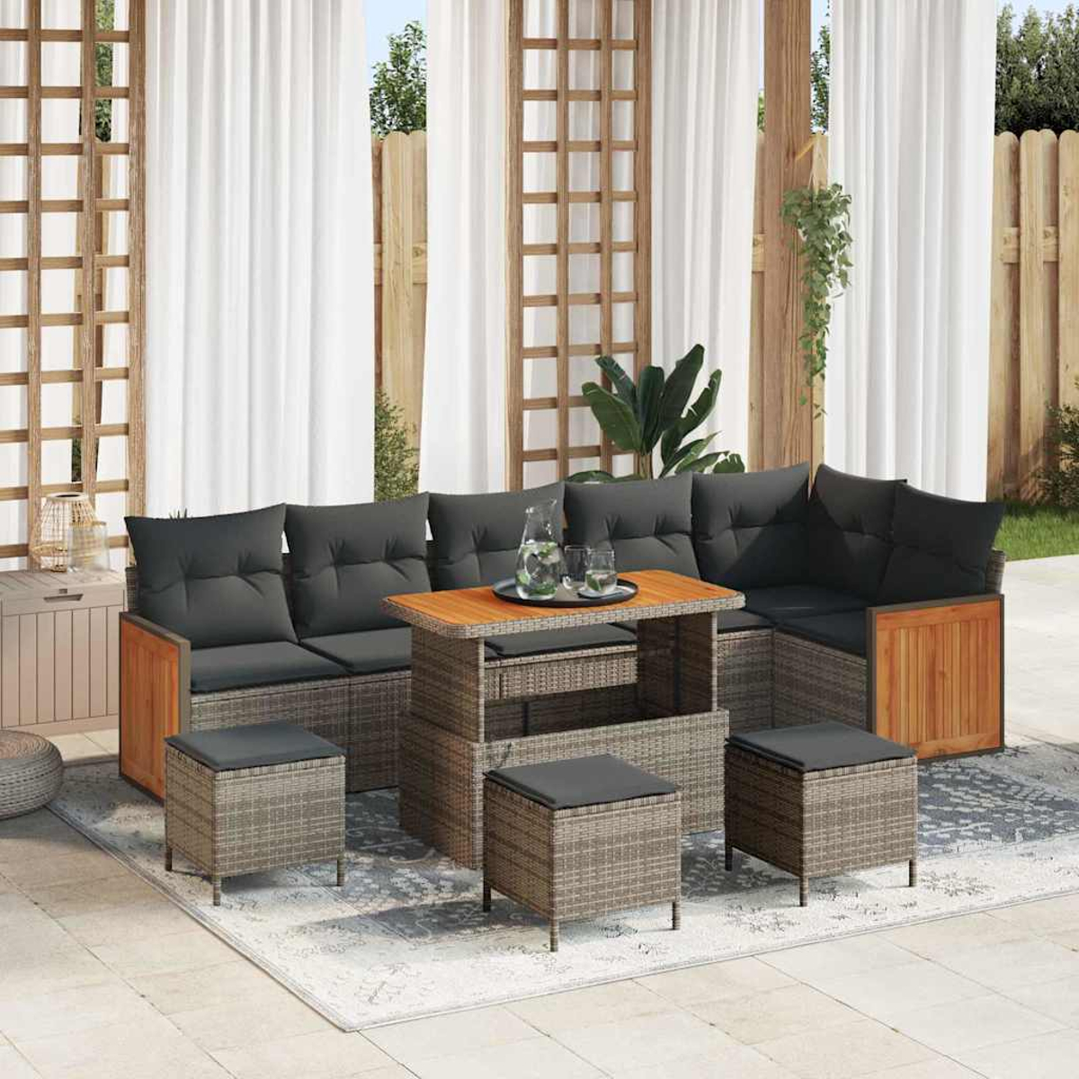 Gartencouch mit grauen Kissen, Holztisch und Rattanhockern auf einem Teppich. Weiße Vorhänge und Holzzaun im Hintergrund.
