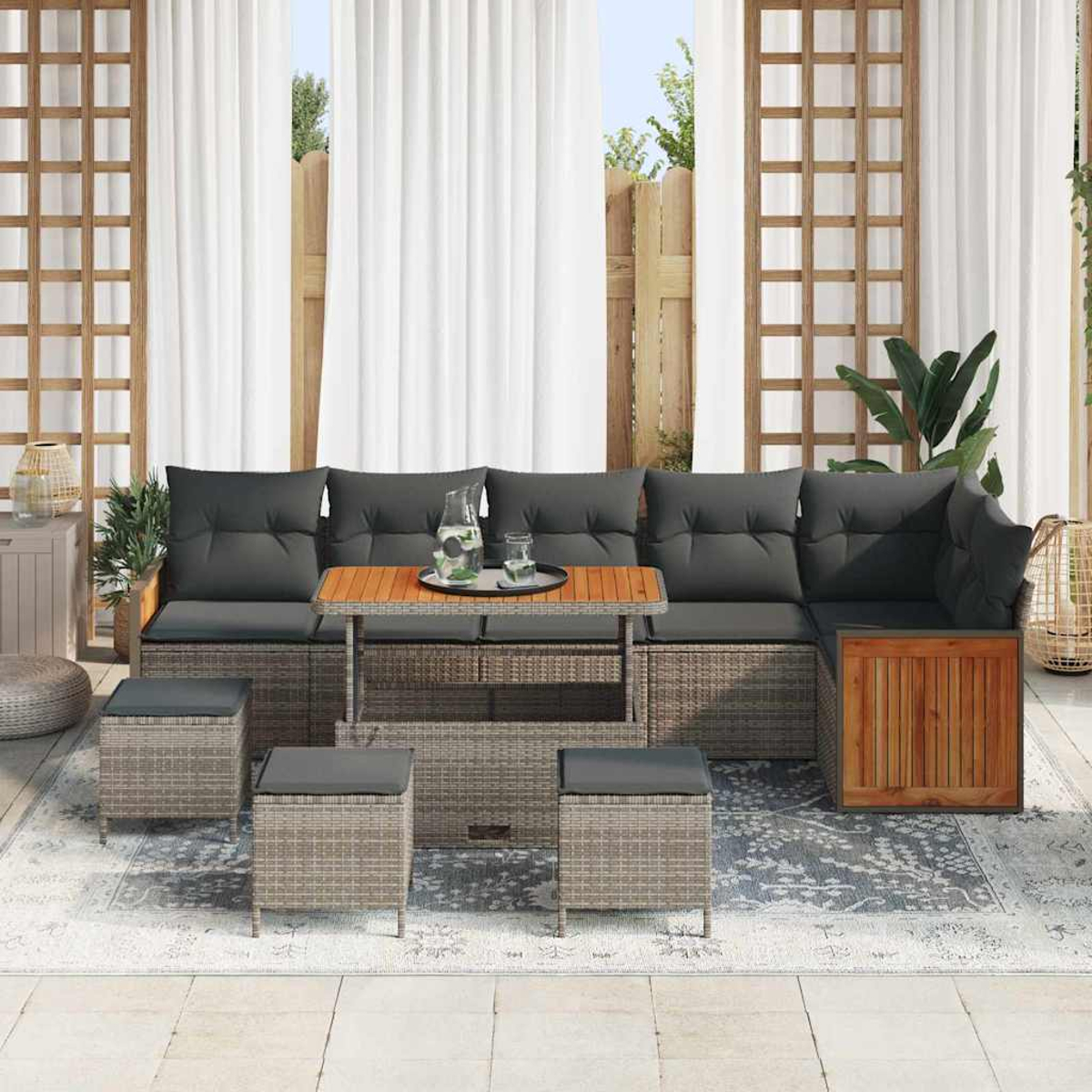 Gartenmöbel-Set mit grauen Kissen, einem Holztisch und Hockern auf einem gemusterten Teppich, in der Nähe weißer Vorhänge und eines Holzzauns.