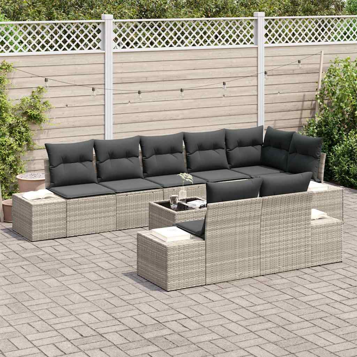Juego de muebles de patio con cojines grises. Incluye un sofá, dos otomanas y una mesa pequeña. El fondo tiene una cerca y plantas.