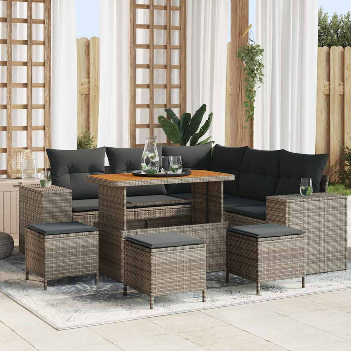 Set di mobili in vimini da esterno con cuscini neri. Include tavolo, divano e pouf. Sfondo con tende bianche e struttura in legno.