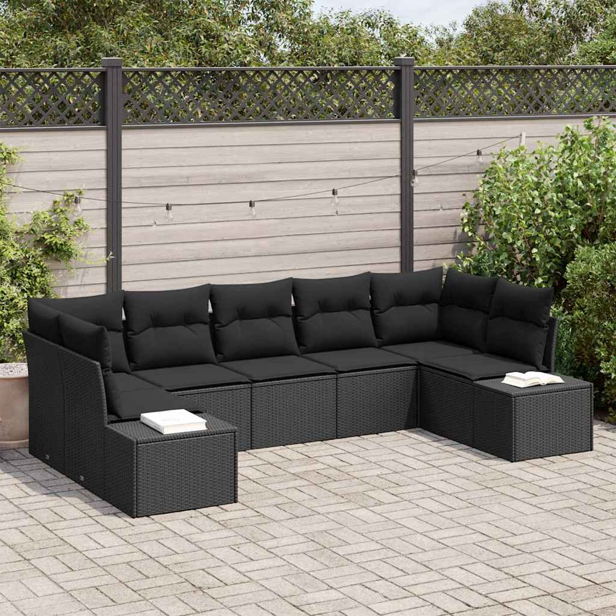 Ein schwarzes Outdoor-Ecksofa mit Kissen steht auf einer Terrasse. Auf dem Hocker liegt ein Buch. Im Hintergrund ein Zaun und Grün.