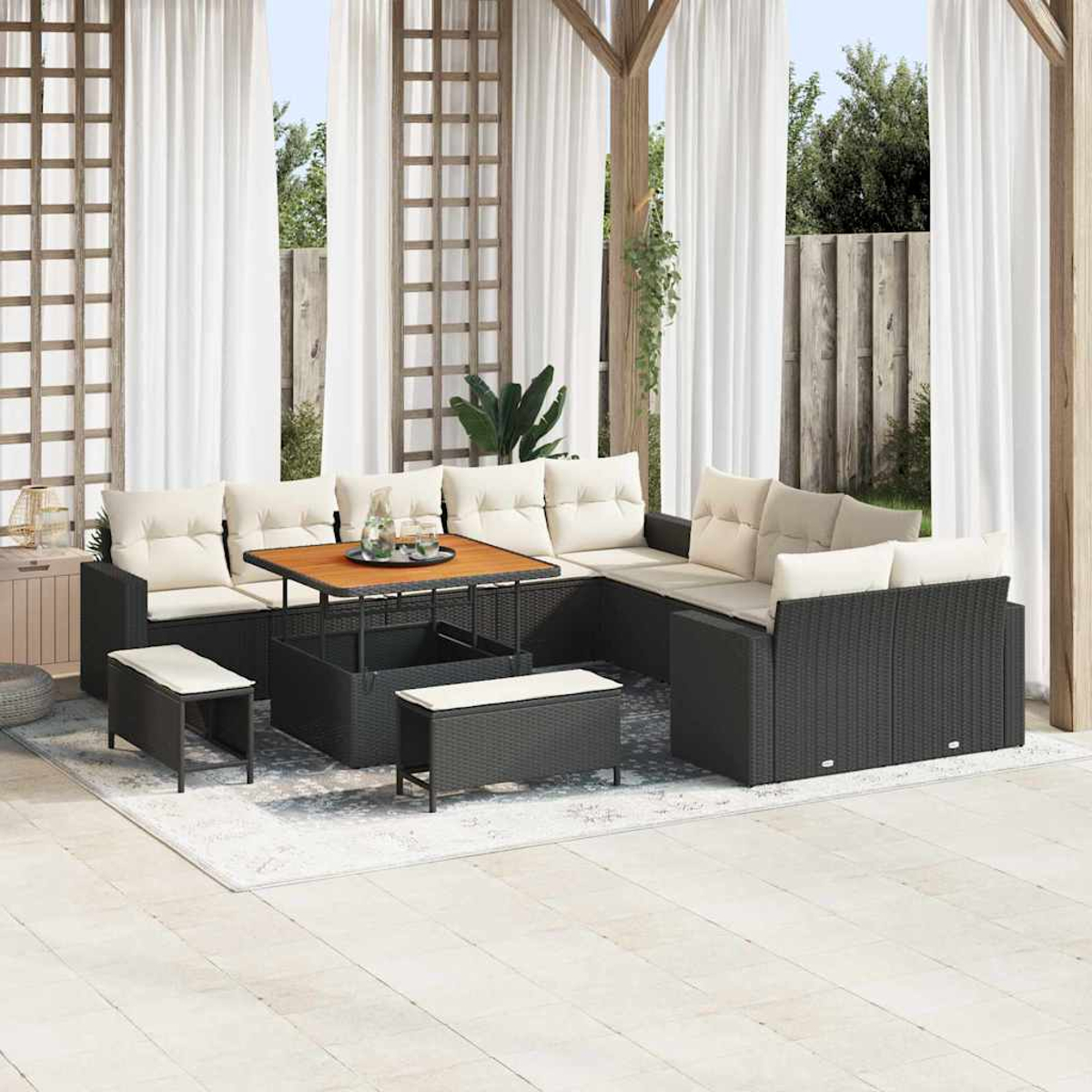 Juego de patio exterior con muebles de mimbre negro, cojines beige, mesa de madera y cortinas blancas. La escena incluye una alfombra y vegetación.