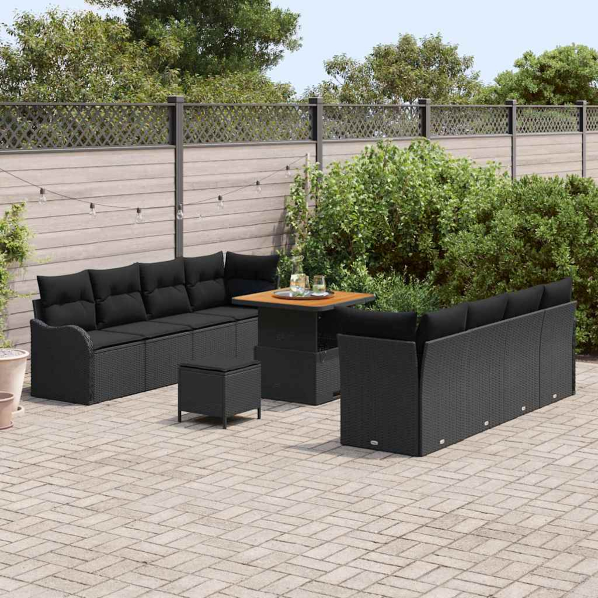 Terrassenmöbel-Set mit schwarzem Korbmöbeln, darunter ein Sofa, Stühle und ein Tisch, auf einer Ziegelterrasse. Grüne Büsche und ein Zaun im Hintergrund.